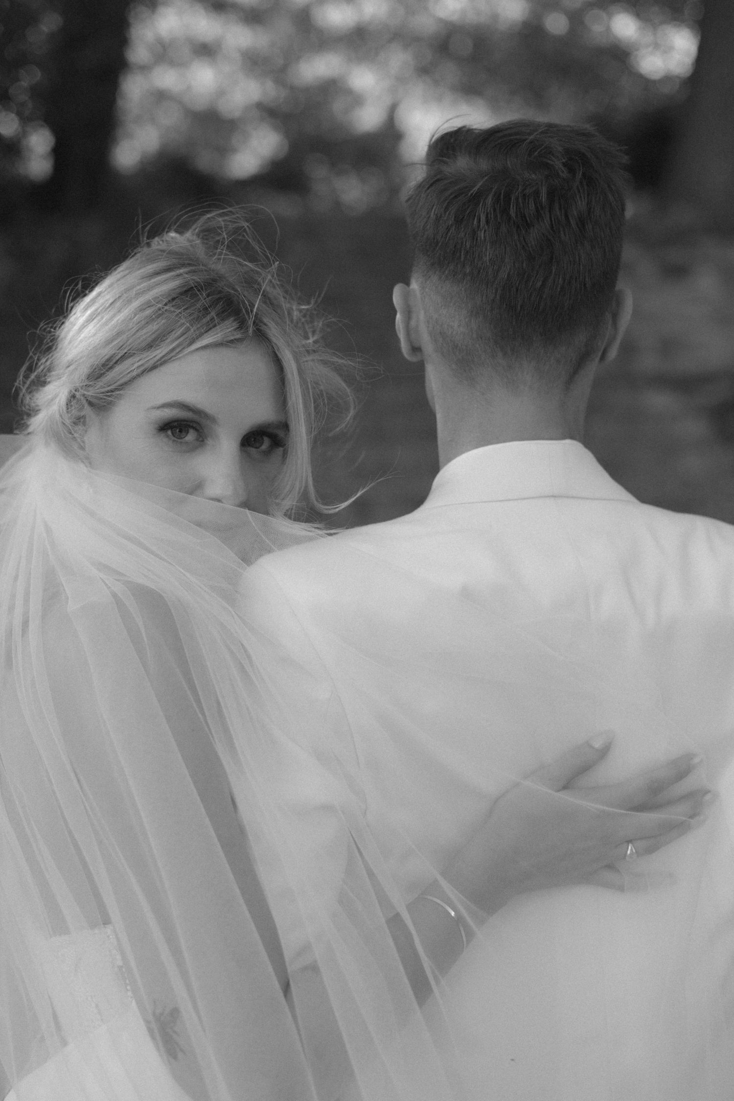 Black and white wedding photo of bride in veil looking back over grooms shoulder outdoors.