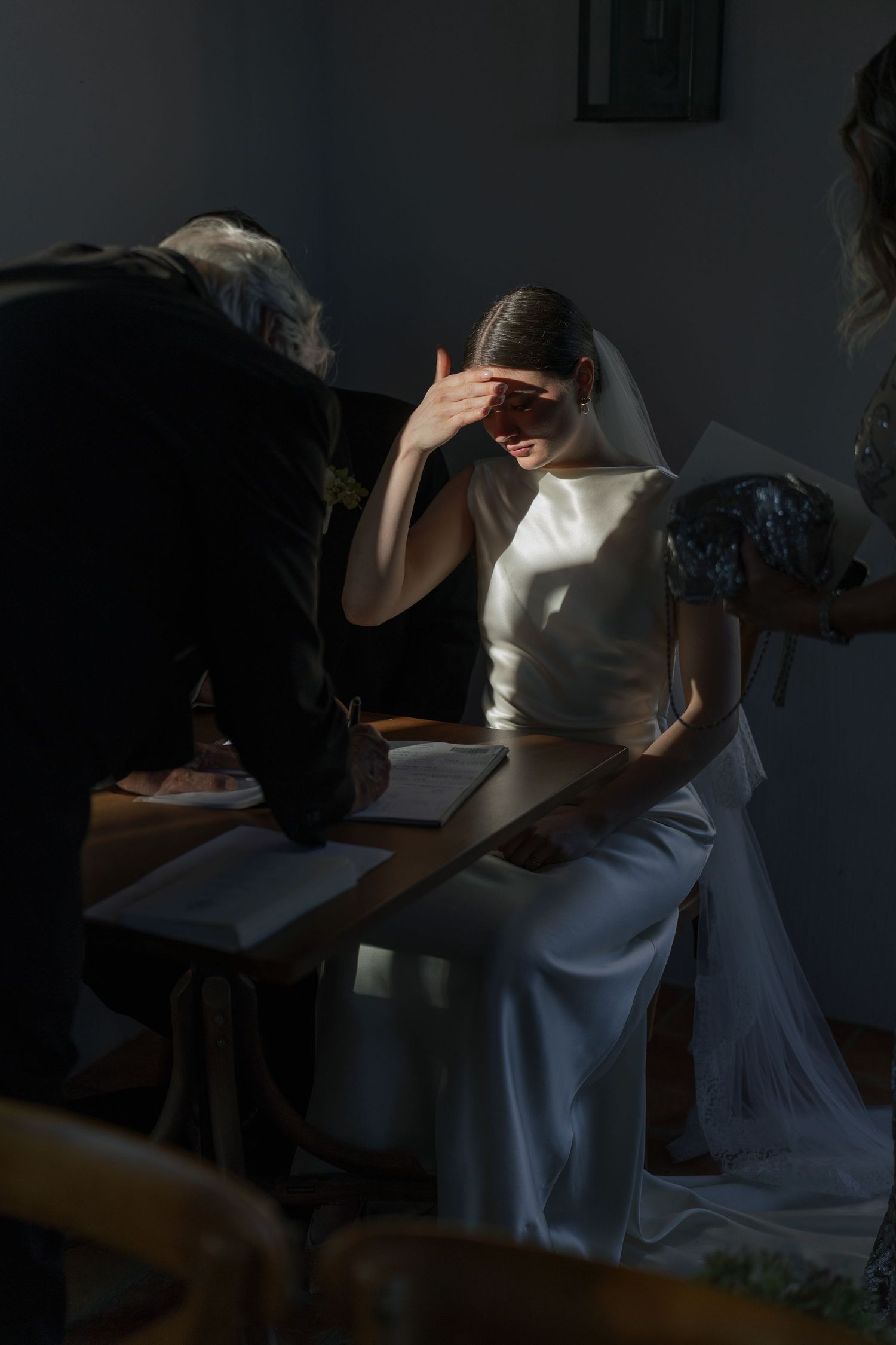 Bride in white gown and veil shielding eyes from dramatic sunlight streaming in during document signing.
