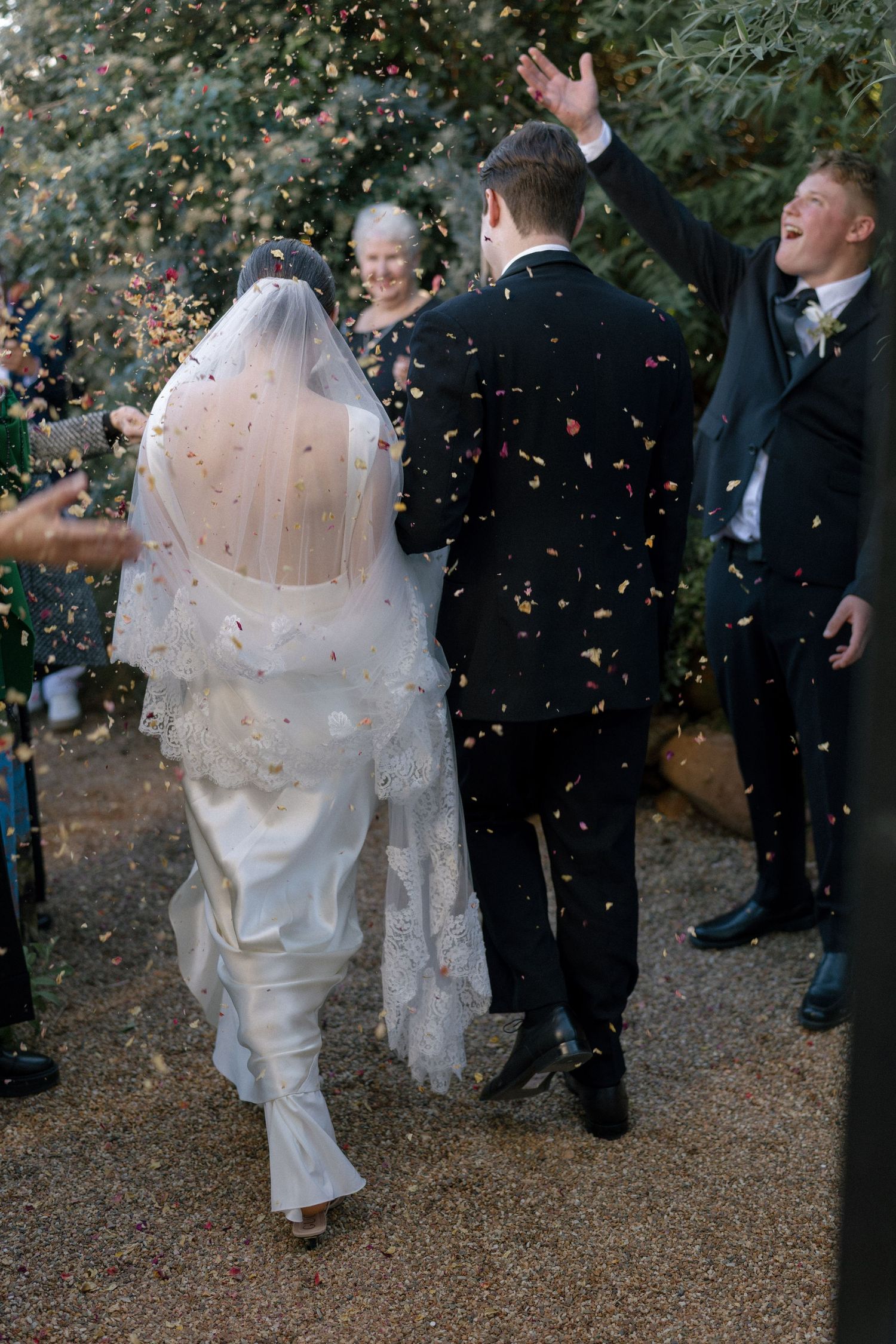 Bride and groom walking away through confetti shower as guests celebrate outside chapel after wedding ceremony.