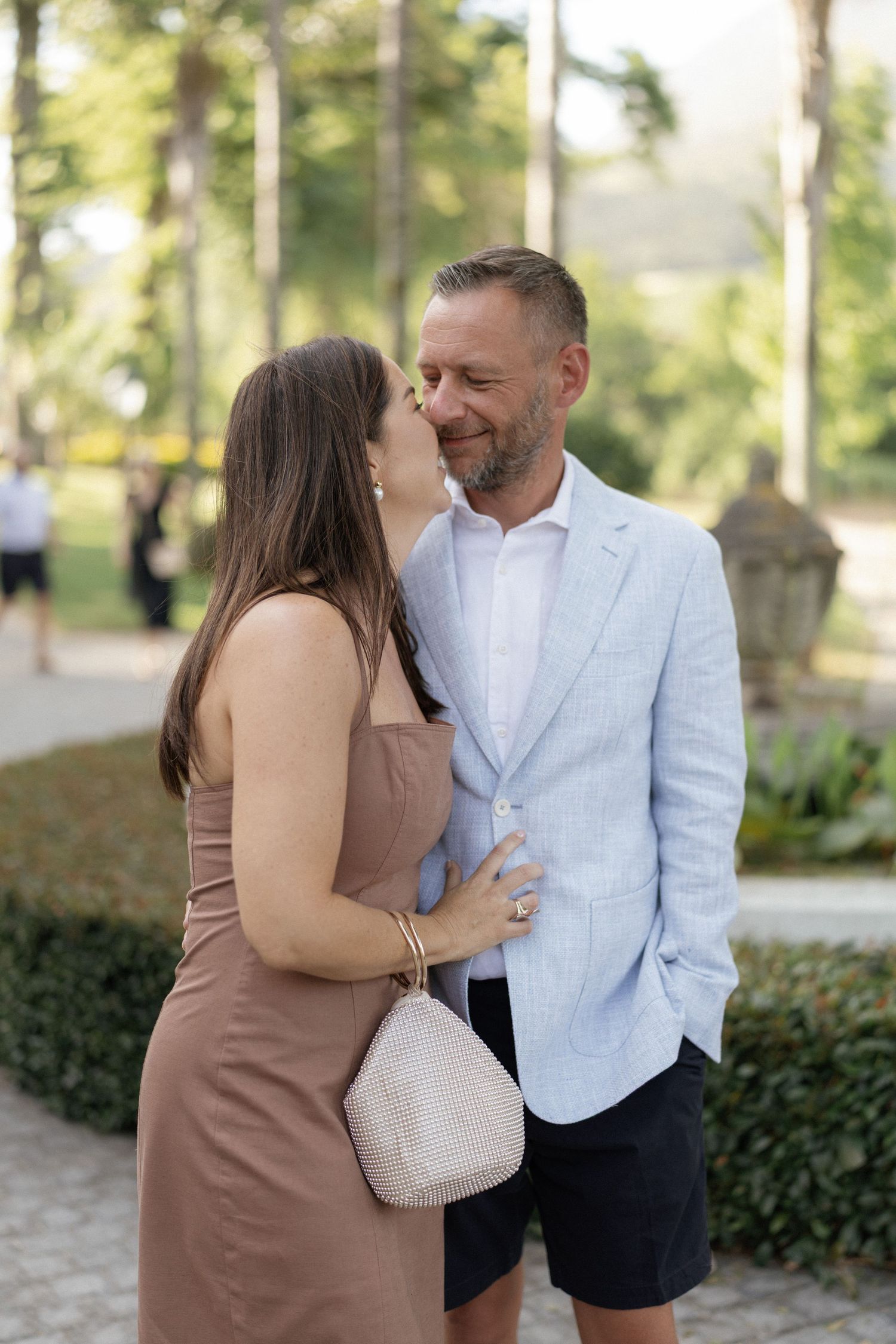 A smiling couple embraces outdoors in a lush garden, woman in tan dress holding white bag, man in light blue blazer.