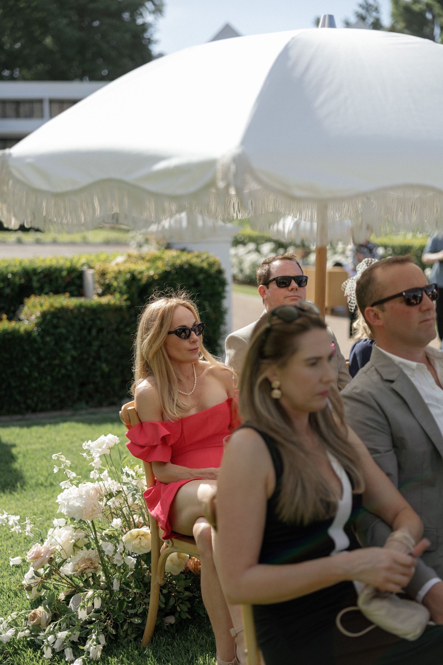 Guests seated outdoors at a summer wedding ceremony under a white fringed umbrella with floral arrangements.