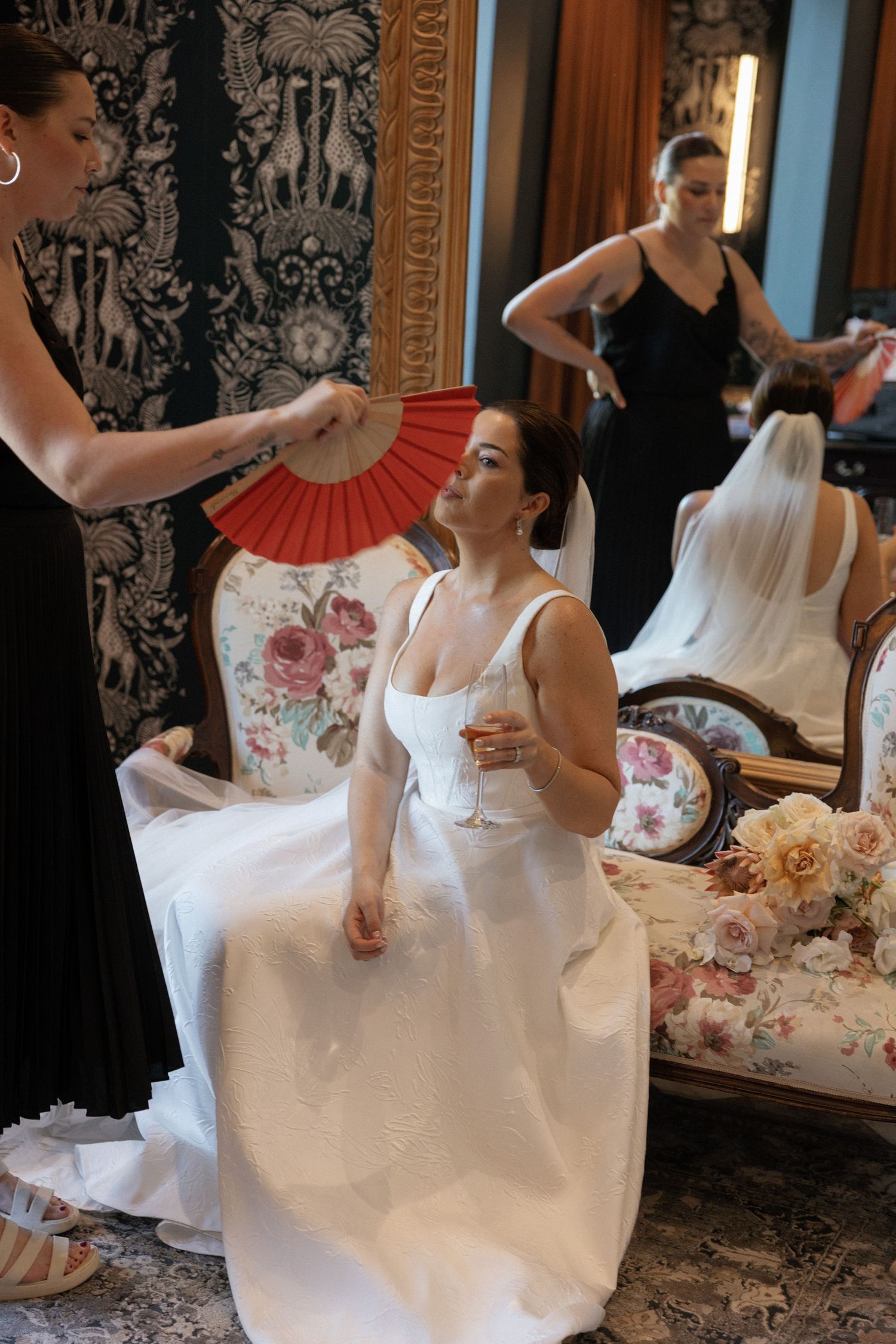 Bride in white gown seated while bridesmaid fans her with red fan in elegant floral room.