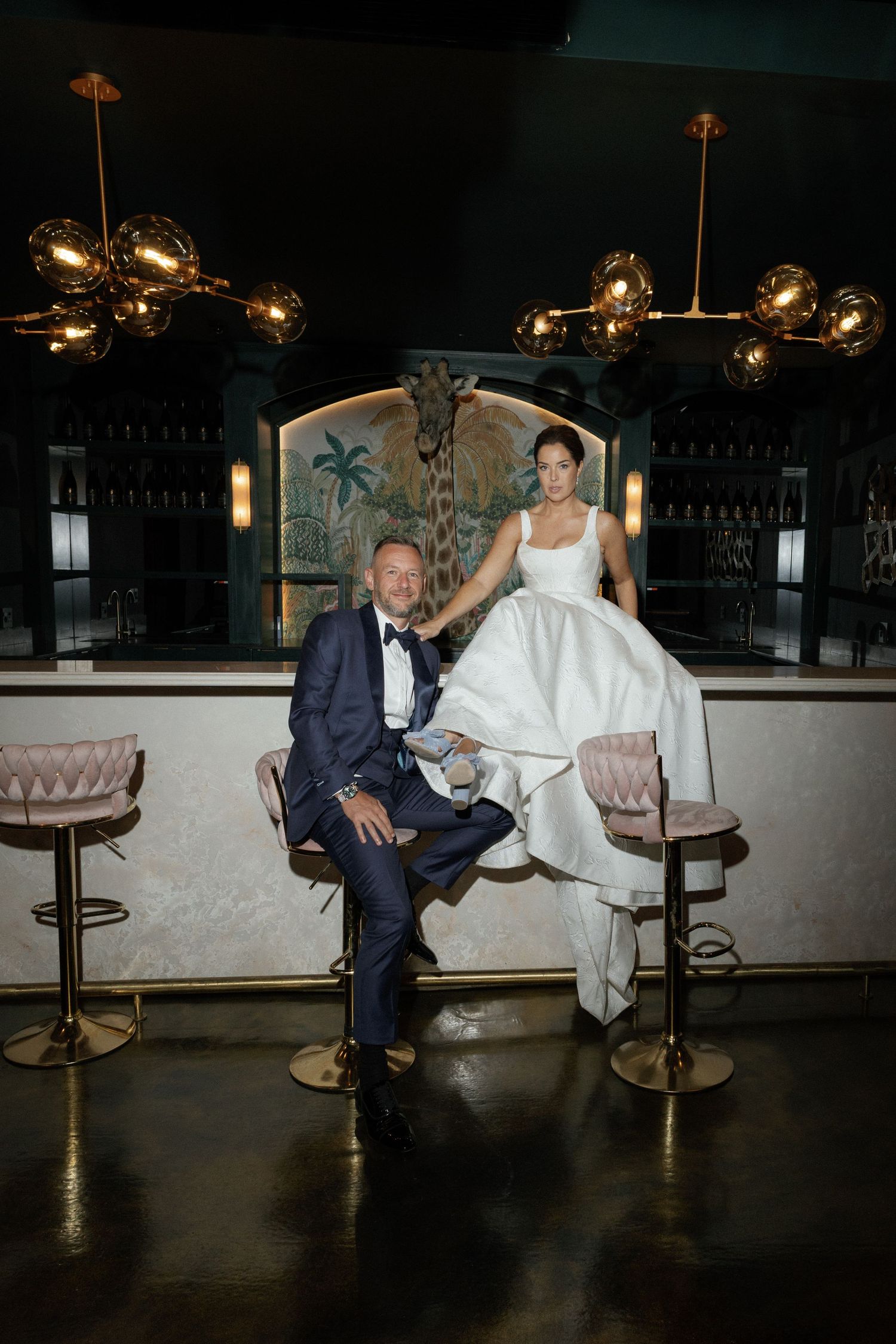 Bride in white gown and groom in navy tuxedo pose elegantly at a stylish bar with gold chandeliers.
