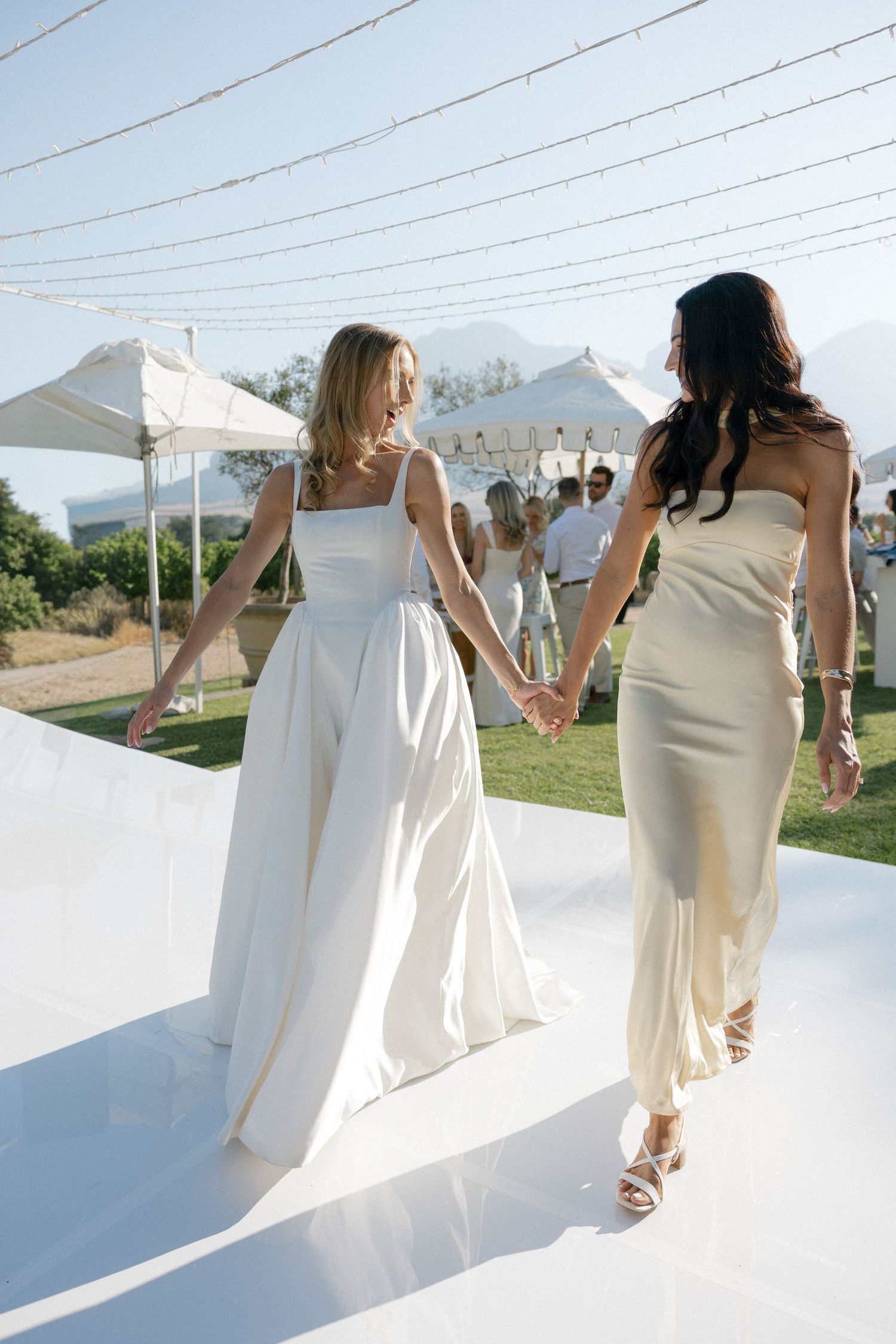 bride in white and guest cream dresses hold hands walking at an outdoor wedding reception under string lights.