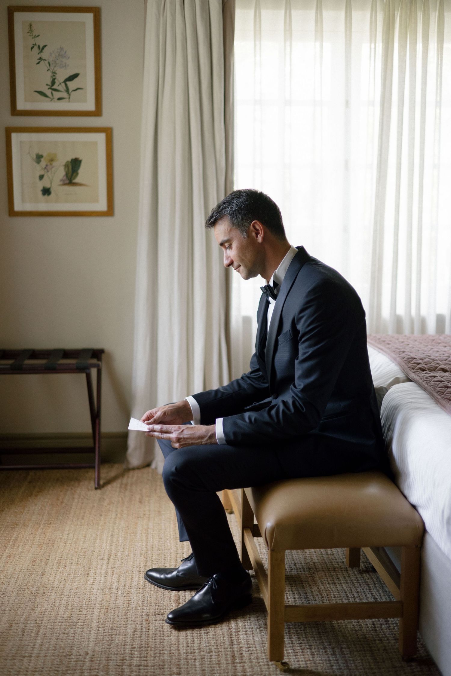 Groom in black tuxedo sitting on bed reading a letter by window light before wedding ceremony.