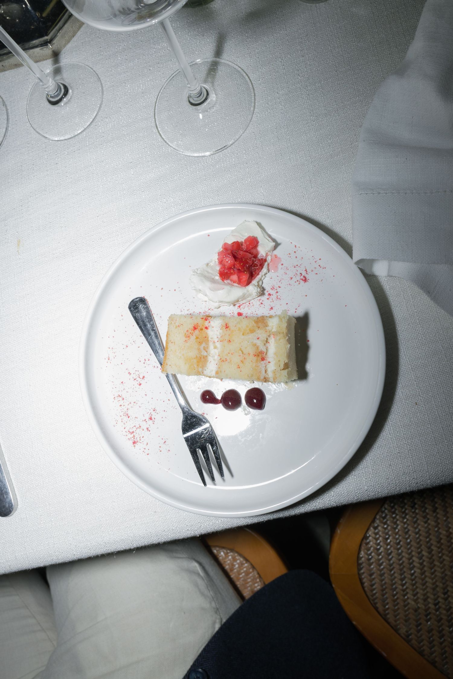 White plate with a slice of white wedding cake, raspberries, cherries, and a fork on a white tablecloth near wine glasses.