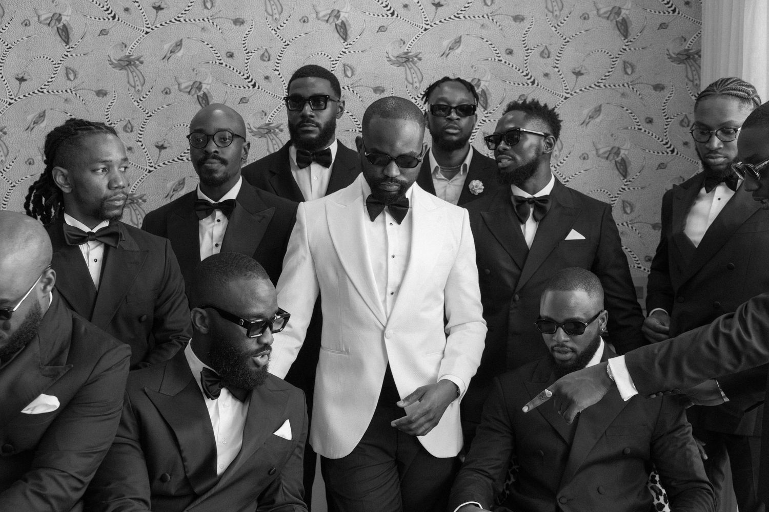 Groom in white tuxedo jacket surrounded by groomsmen in black suits, elegant black and white wedding photo.