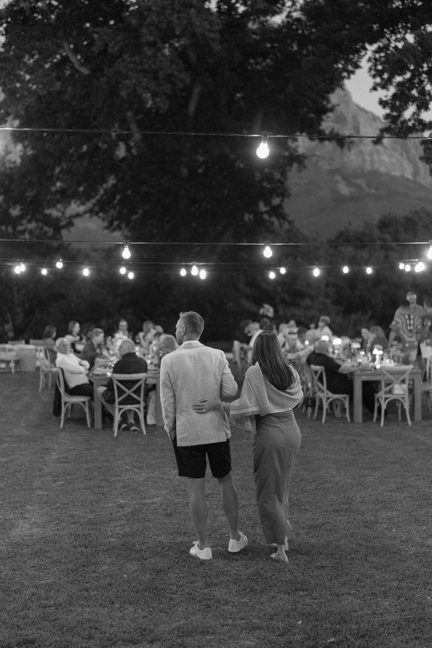 Black and white photo of a couple embracing at an outdoor evening garden party with string lights and guests dining.