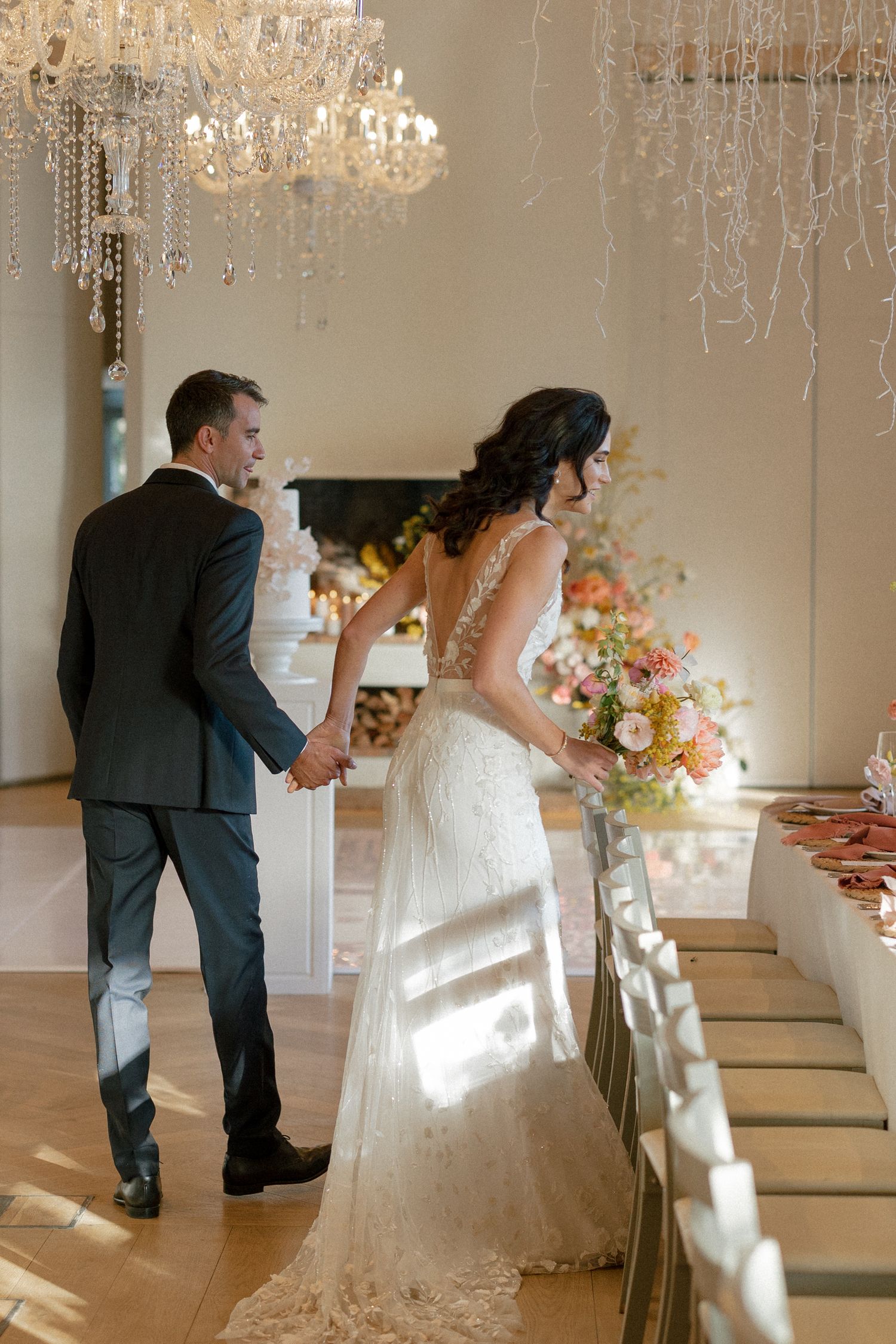Bride and groom holding hands beside long reception table under crystal chandeliers.