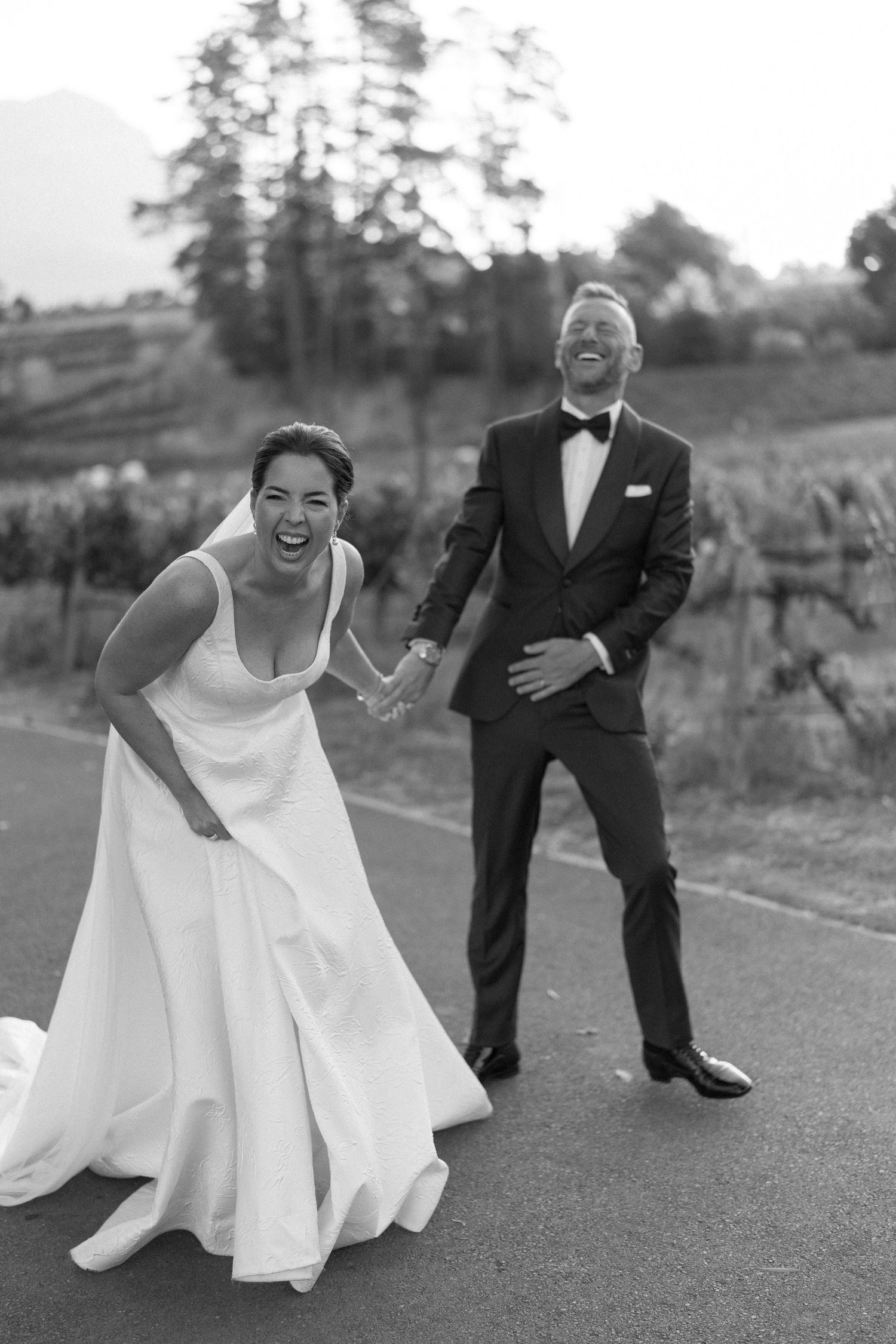 Black and white photo of laughing bride in white gown and groom in tuxedo holding hands on a road.
