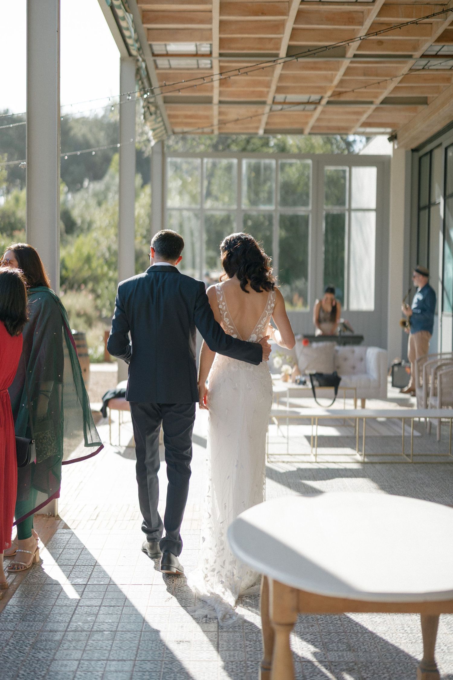 Bride in backless lace gown walks with groom through elegant outdoor wedding venue corridor.