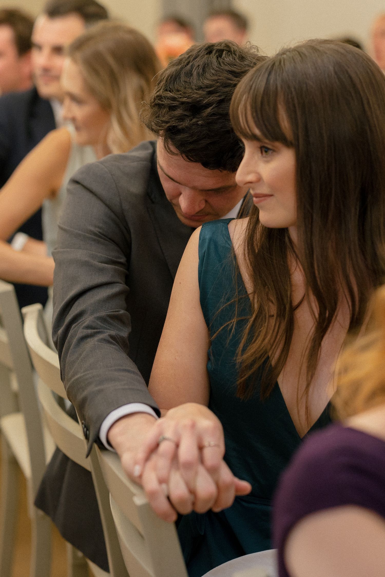 Wedding guests holding hands tenderly during the ceremony seated in white chairs.