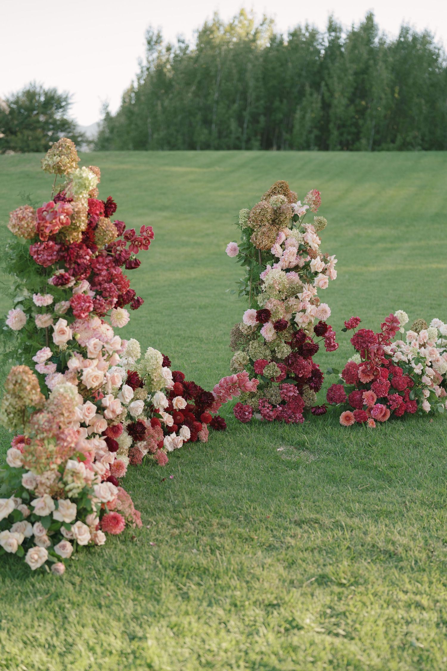 Lush floral arrangements with pink, red, and cream roses and hydrangeas on green lawn at outdoor wedding ceremony.