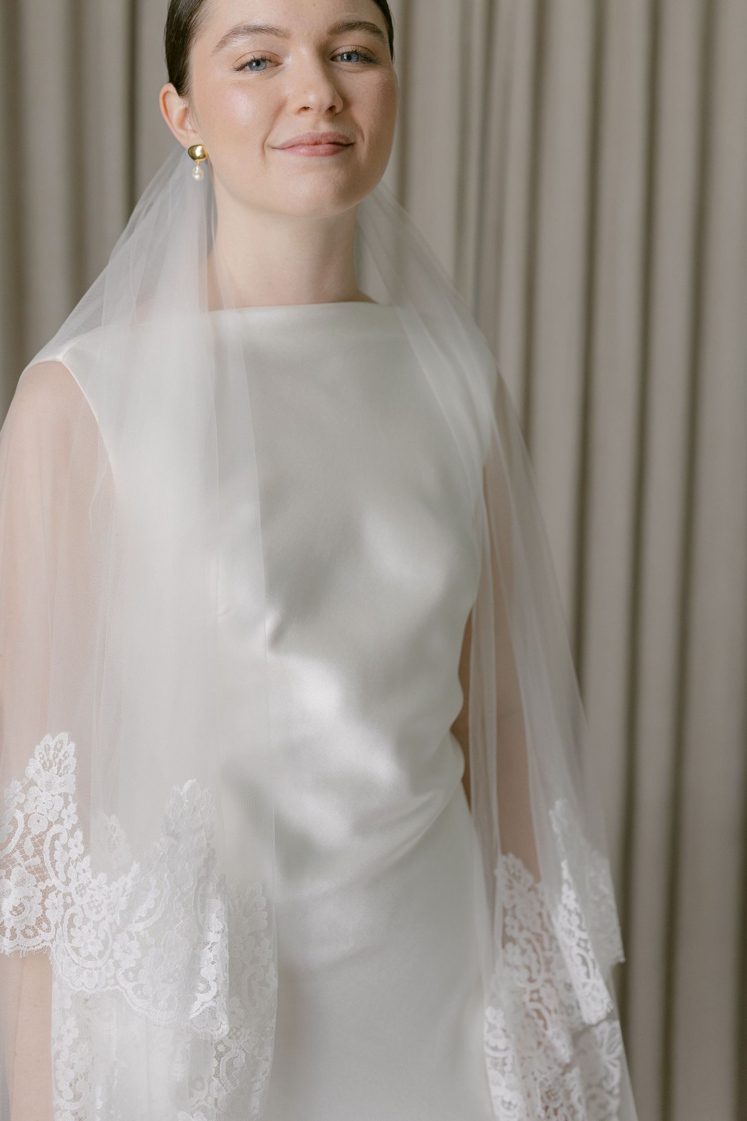 Bride in a sleek white gown and lace-trimmed veil smiling softly in a neutral interior.