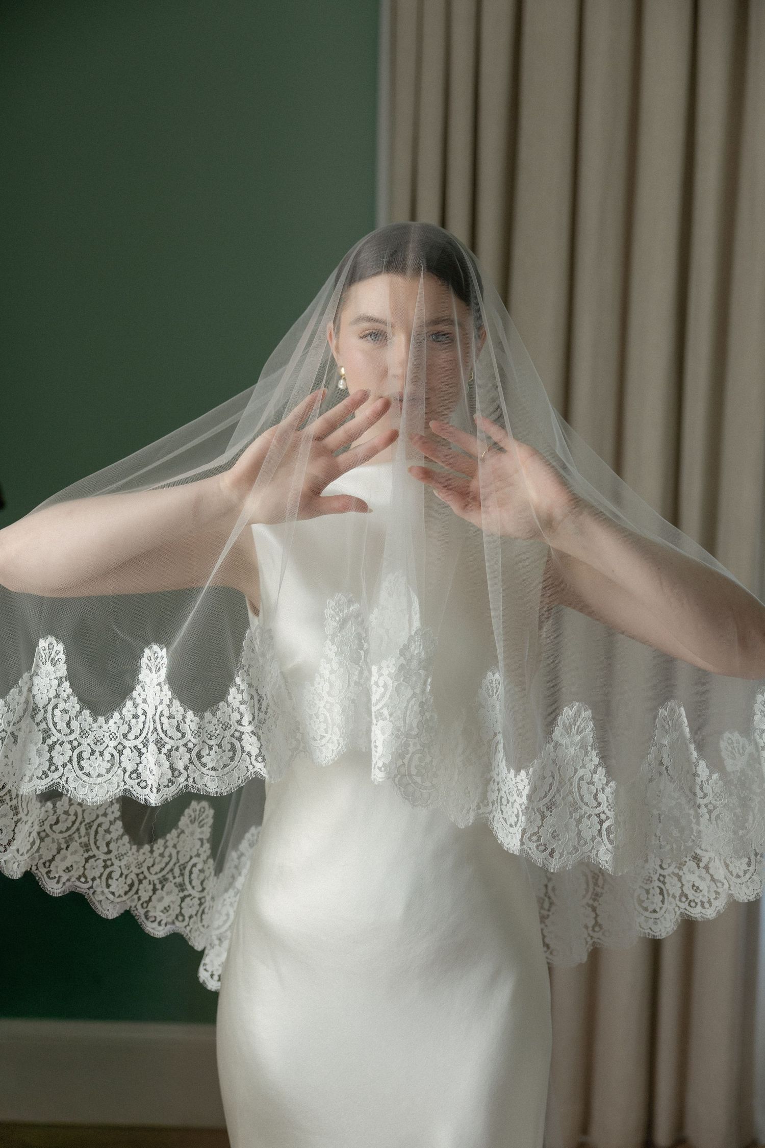 Bride in satin gown lifts lace-edged veil with both hands raised to face.