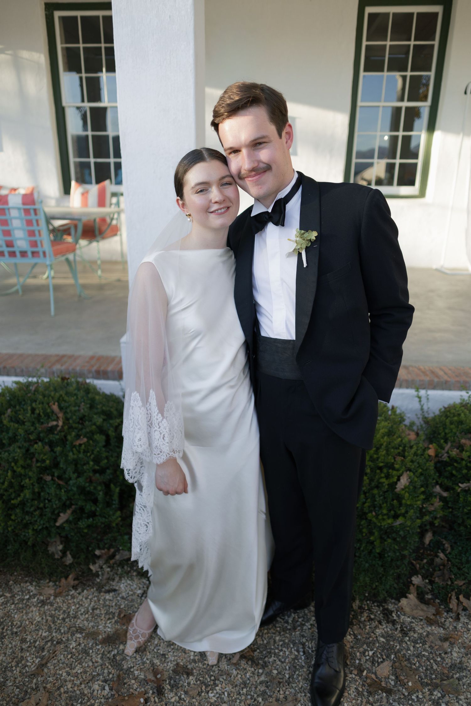 Bride in elegant white gown and groom in black tuxedo posing together outside a white manor house.
