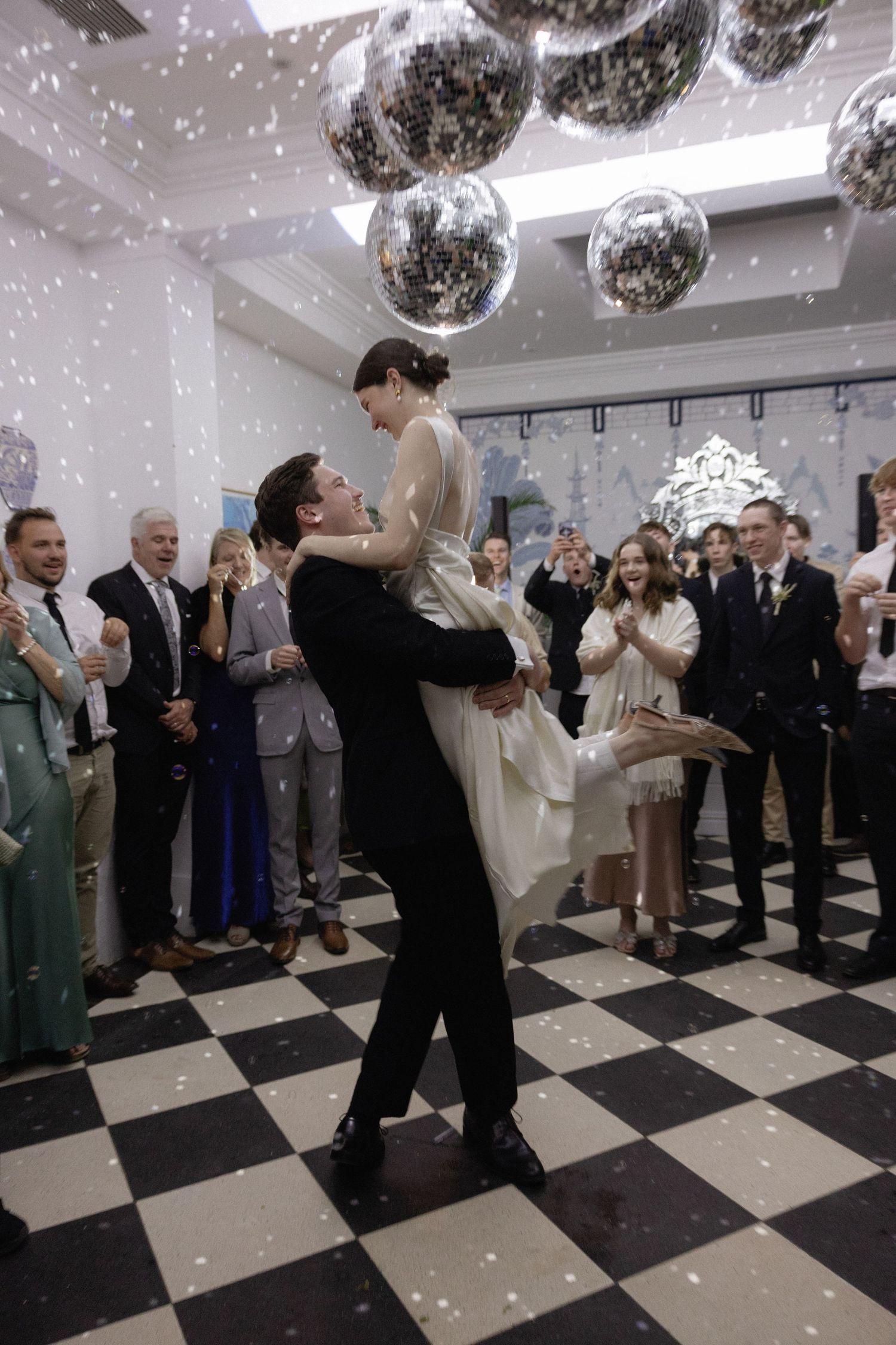 Groom lifts bride on checkered dance floor surrounded by guests under disco balls at elegant wedding reception.