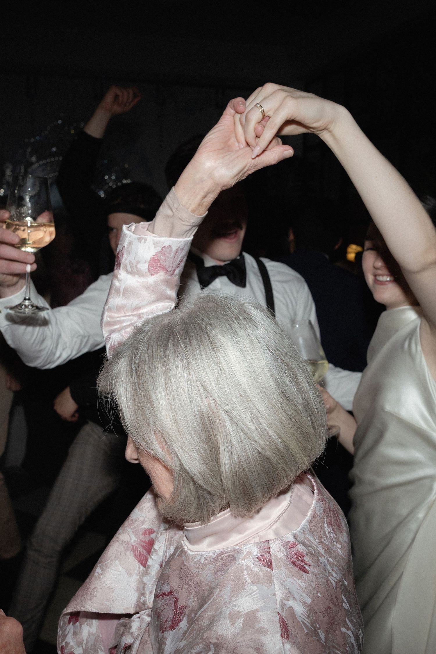 Elderly woman with silver bob hair dancing at a wedding event, holding hands up with other guests.