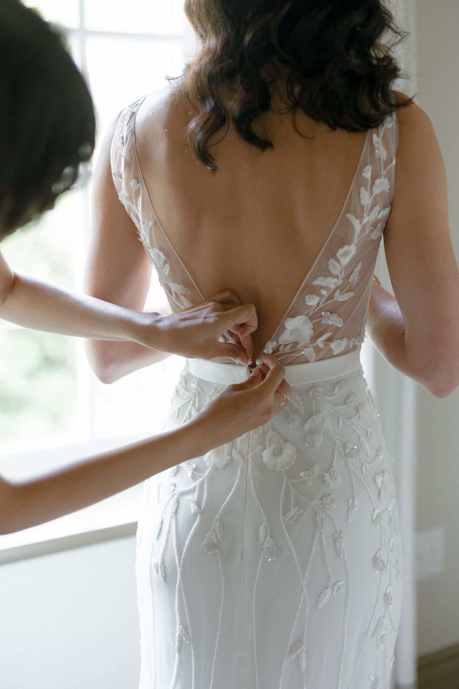 Bridesmaid fastening buttons on back of brides floral embroidered wedding gown near window.