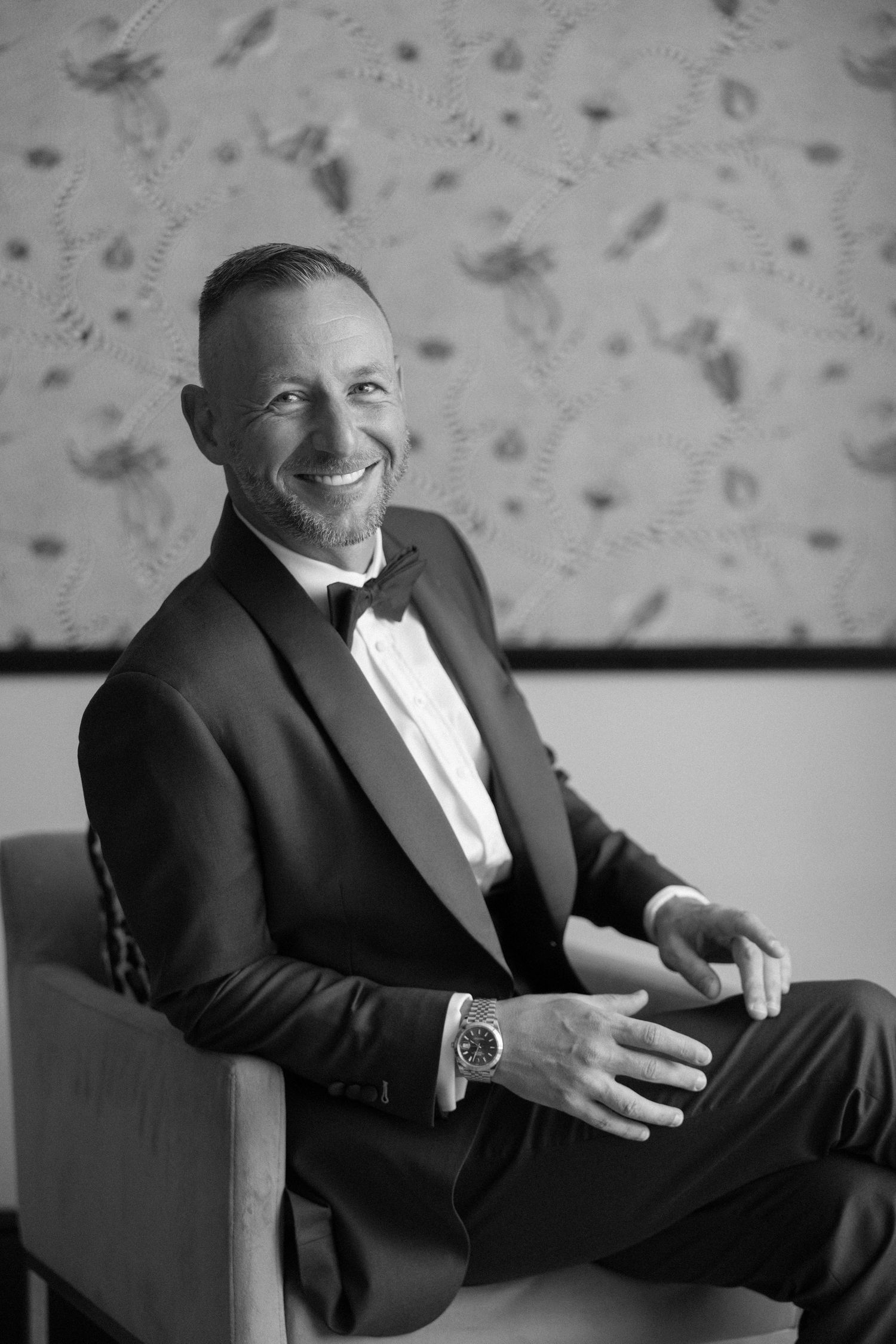 Black and white photo of a smiling man in a tuxedo with bow tie, seated in a chair, wearing a watch.