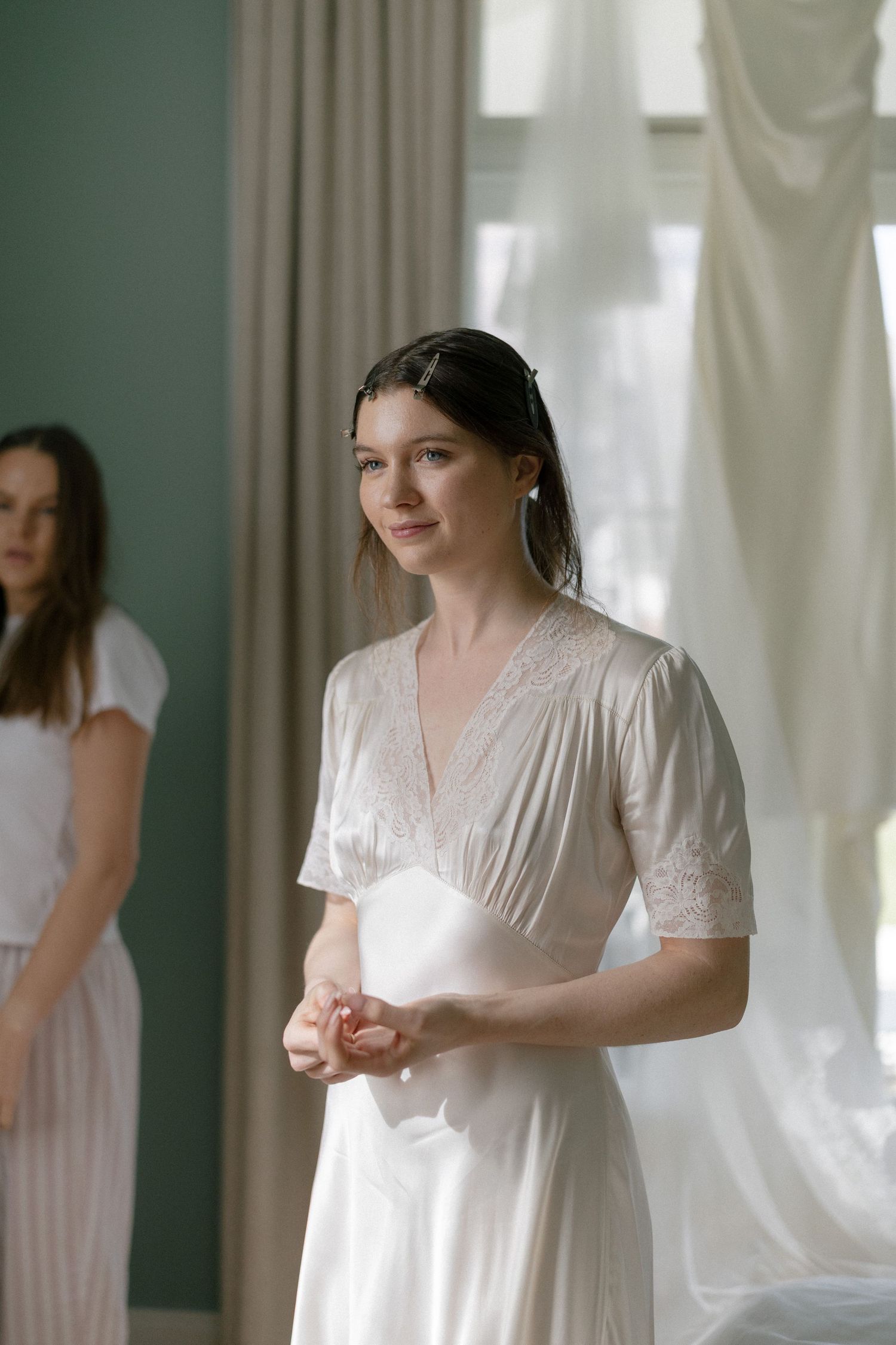 bride in white satin lace dress standing by sheer curtains, with another woman visible in background.