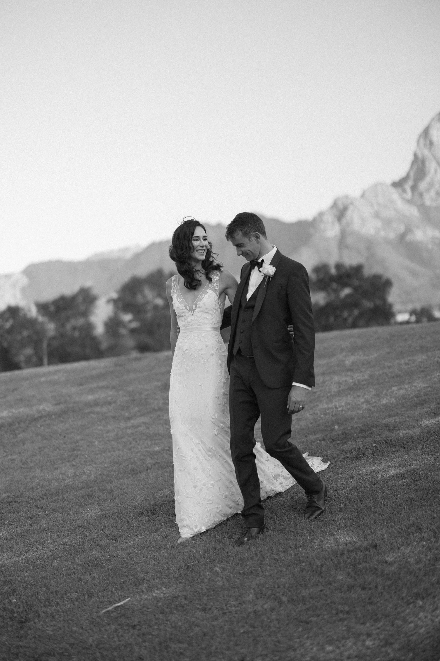 Black and white photo of newlyweds walking and laughing on grassy field with mountains.