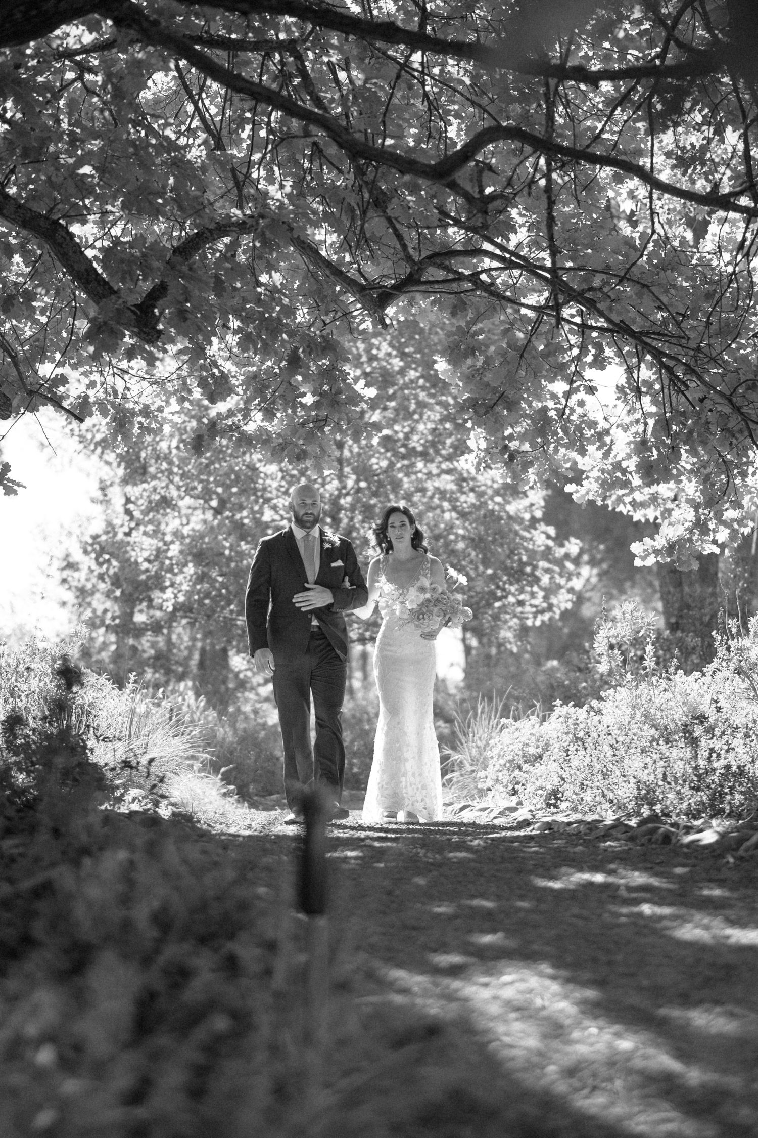 Black and white photo of bride and groom walking hand in hand along a sunlit tree-canopied garden path.