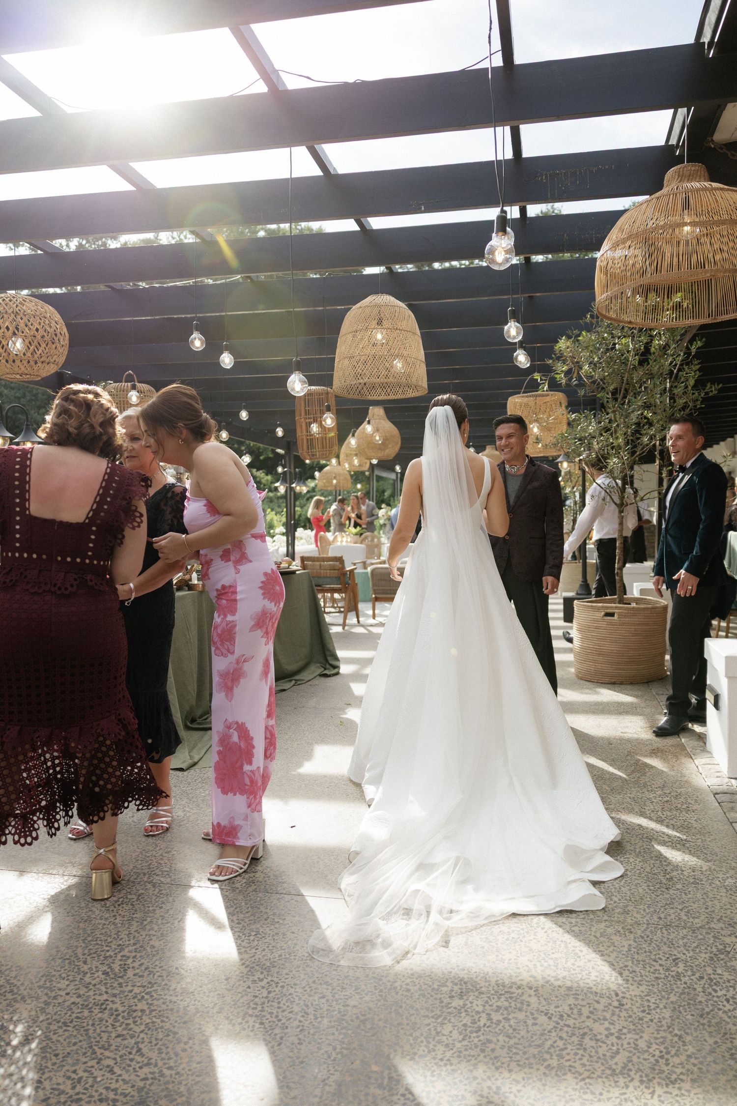 Bride in white gown and veil walks through elegant wedding venue with wicker pendant lights and guests.
