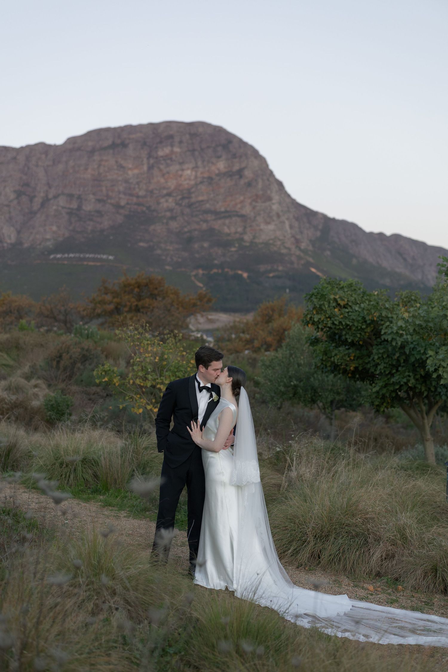 Bride and groom share a romantic kiss on a mountain trail, bride wearing a flowing white gown with long veil.