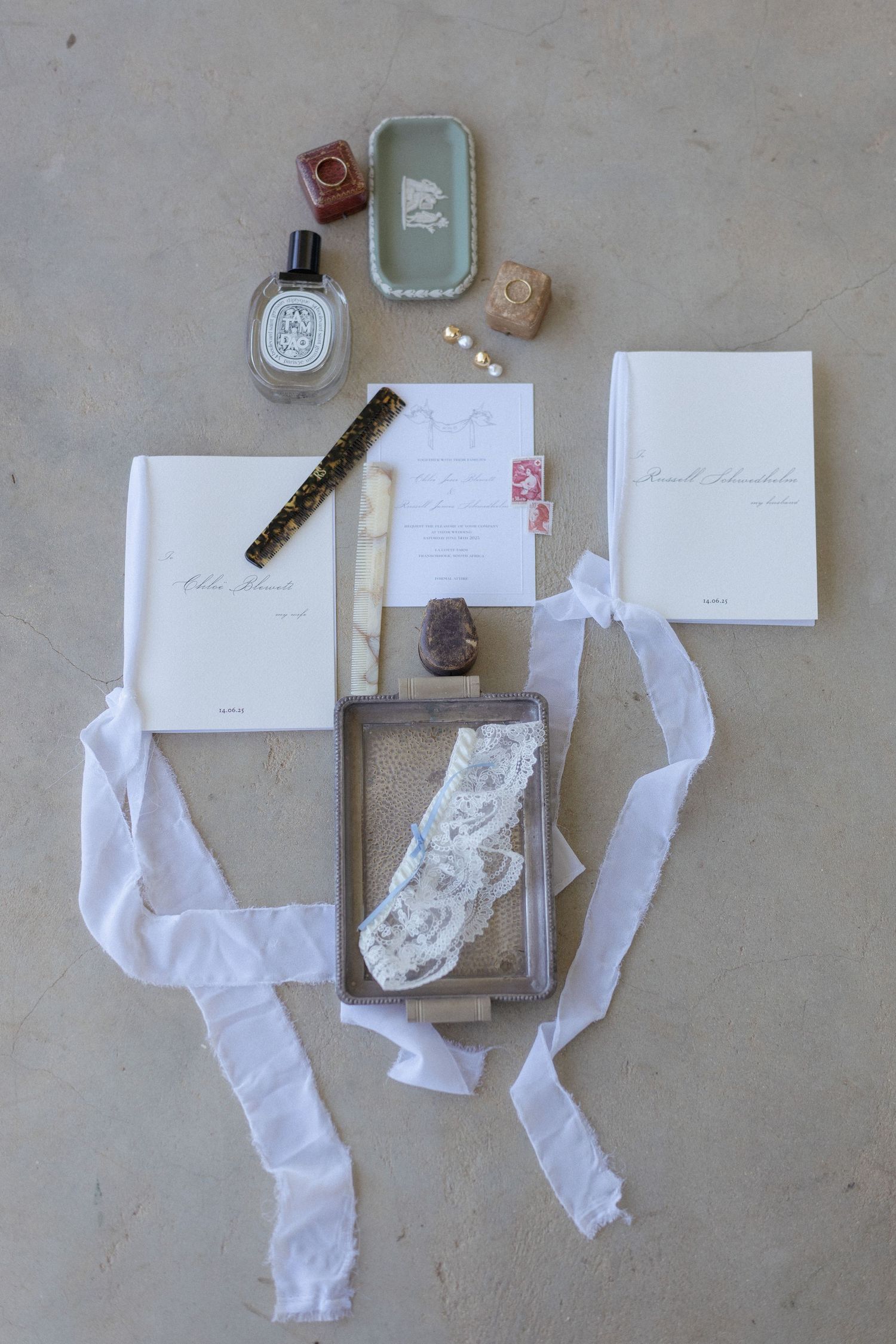 Wedding flat lay with vow books, lace garter, invitation, perfume, and white silk ribbon.