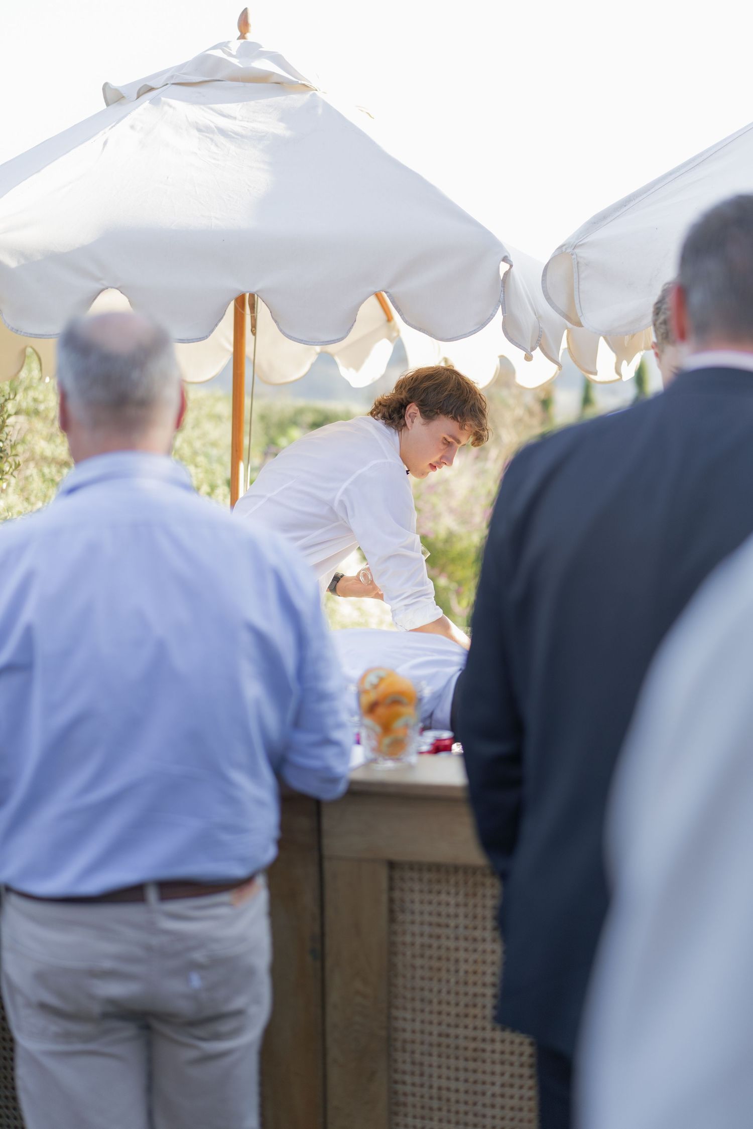 Bartender in white shirt serves guests at outdoor wooden bar station shaded by white scalloped market umbrellas.