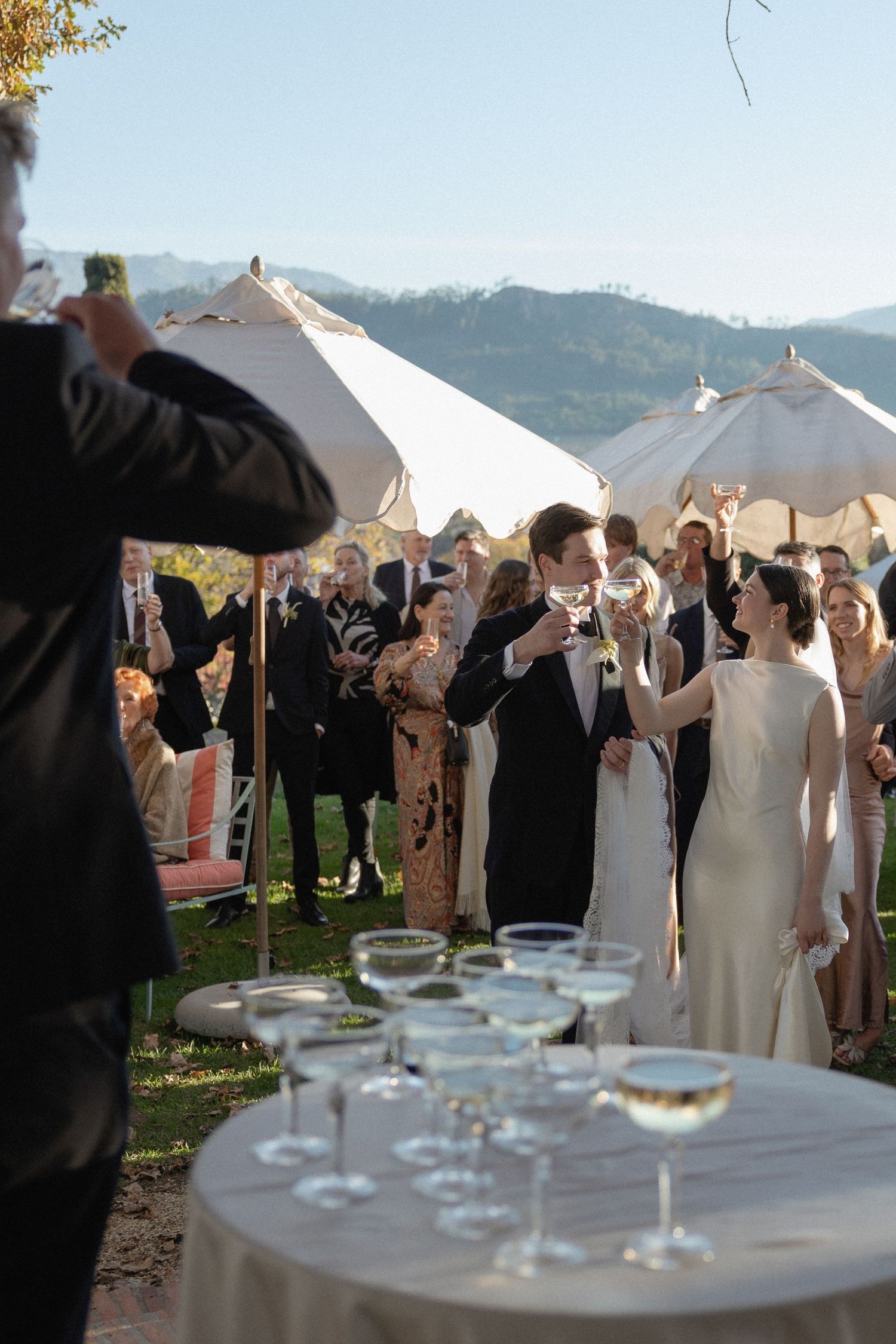 Wedding couple toasting with champagne near a glass tower at outdoor mountain view reception with guests.