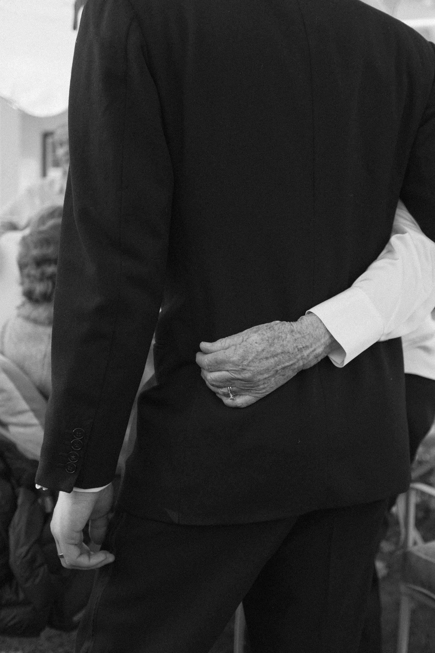 Black and white photo of elderly hands clasped behind a man in a dark suit at a wedding.