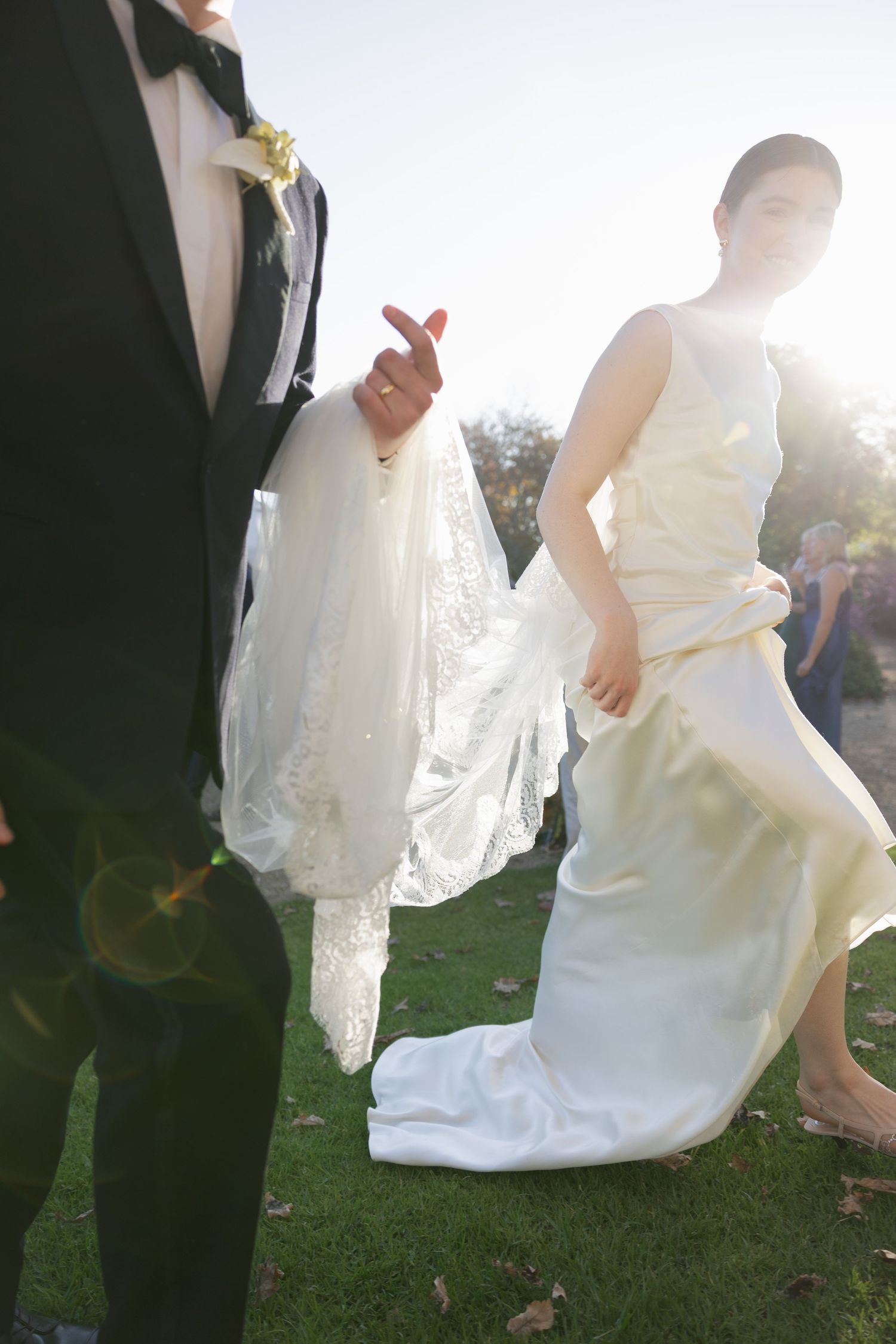 Bride in flowing white gown with long veil walks beside groom in black tuxedo on green grass in sunlight.