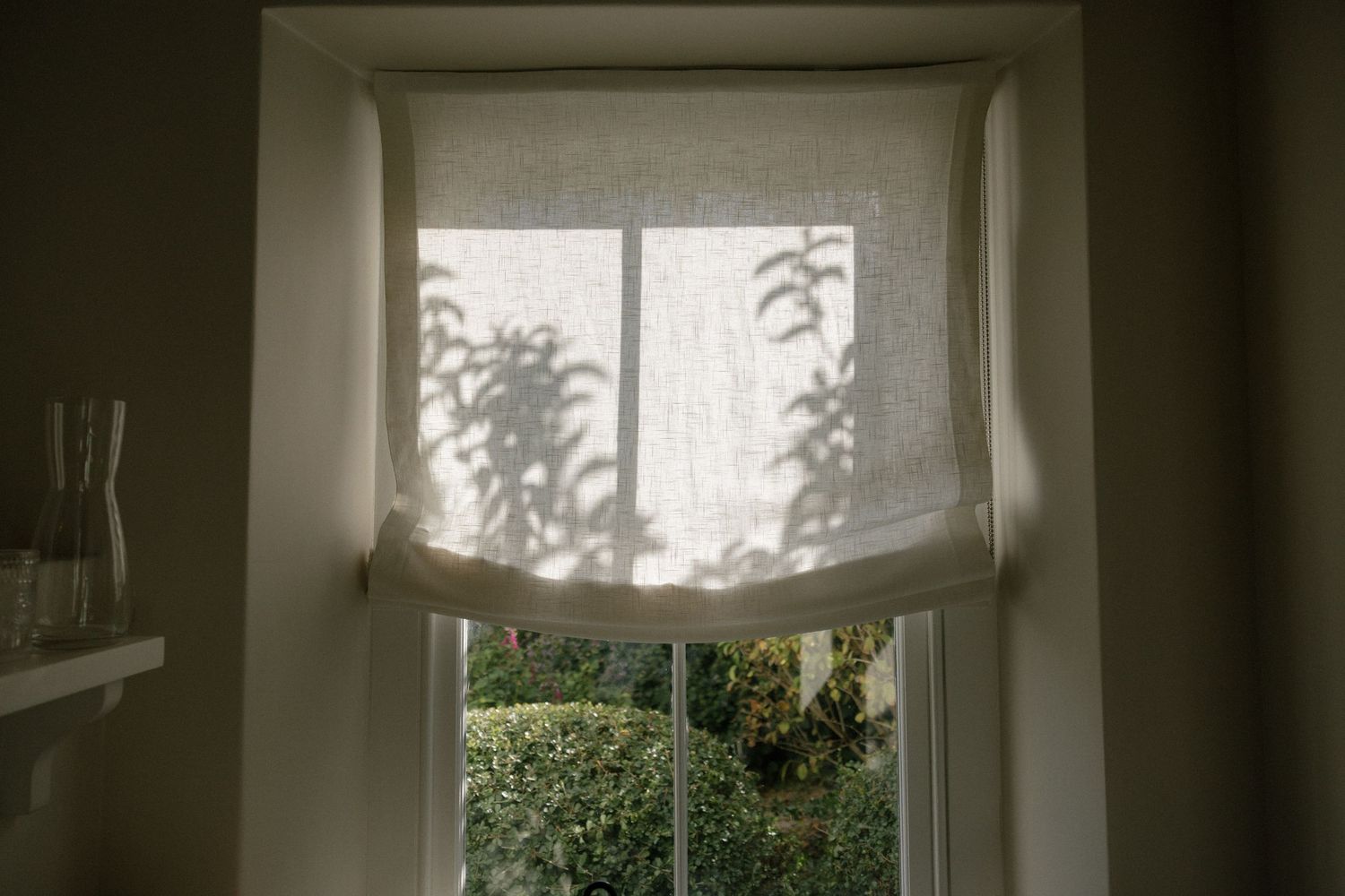 Soft natural light filters through a linen roman shade window with garden greenery visible outside.