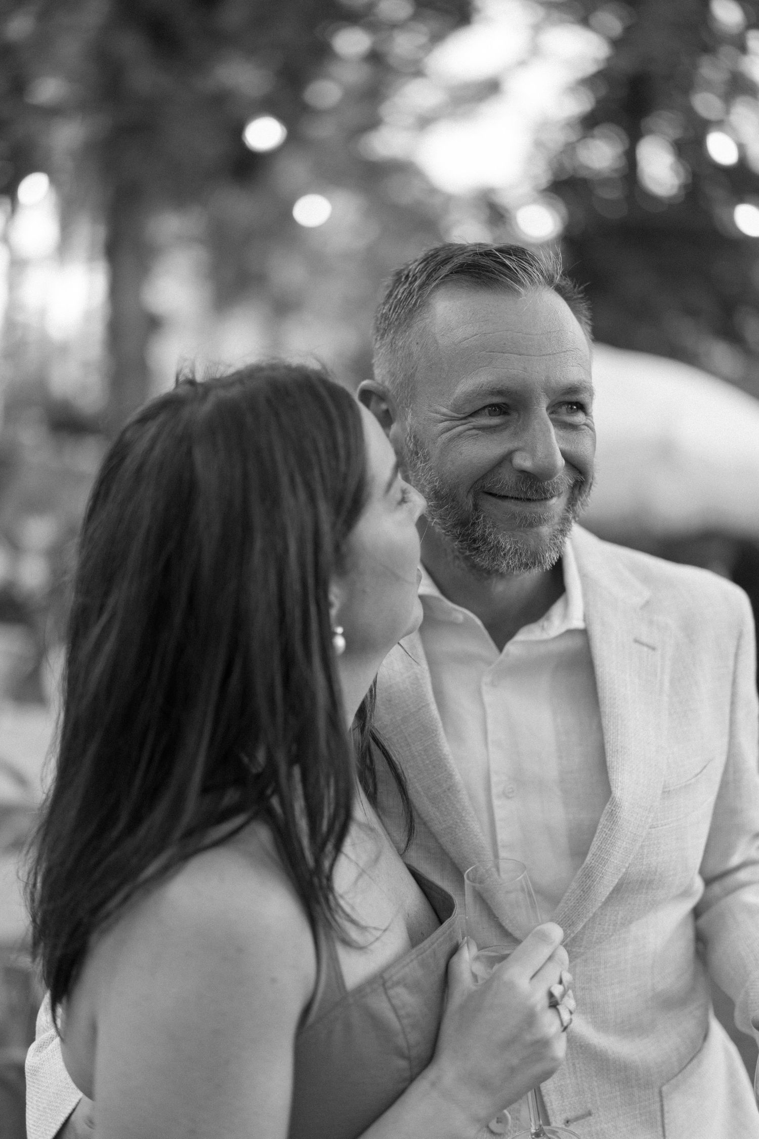Black and white photo of a smiling bearded man in a suit with a woman holding his lapel at an outdoor event.