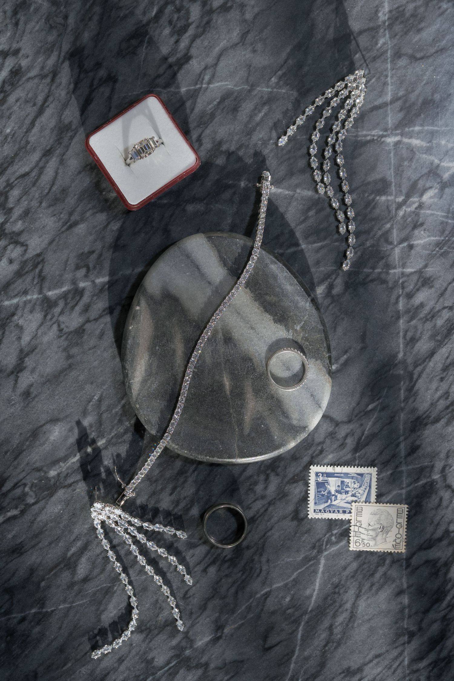 Silver wedding jewellery, ring in red box, chains, and stamps arranged on gray marble surface.