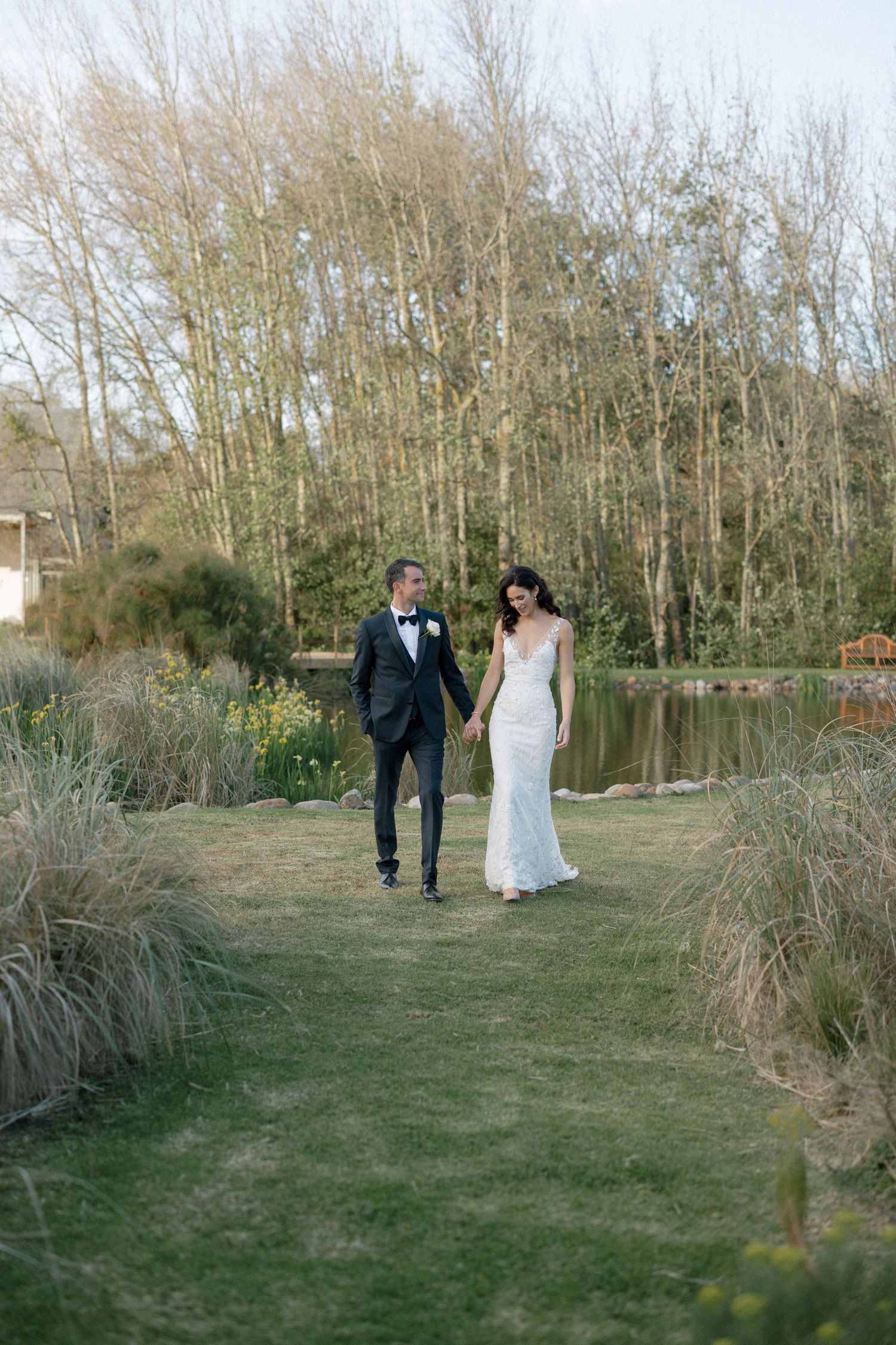 Elegant couple in wedding attire strolling hand in hand along green path beside pond and trees.