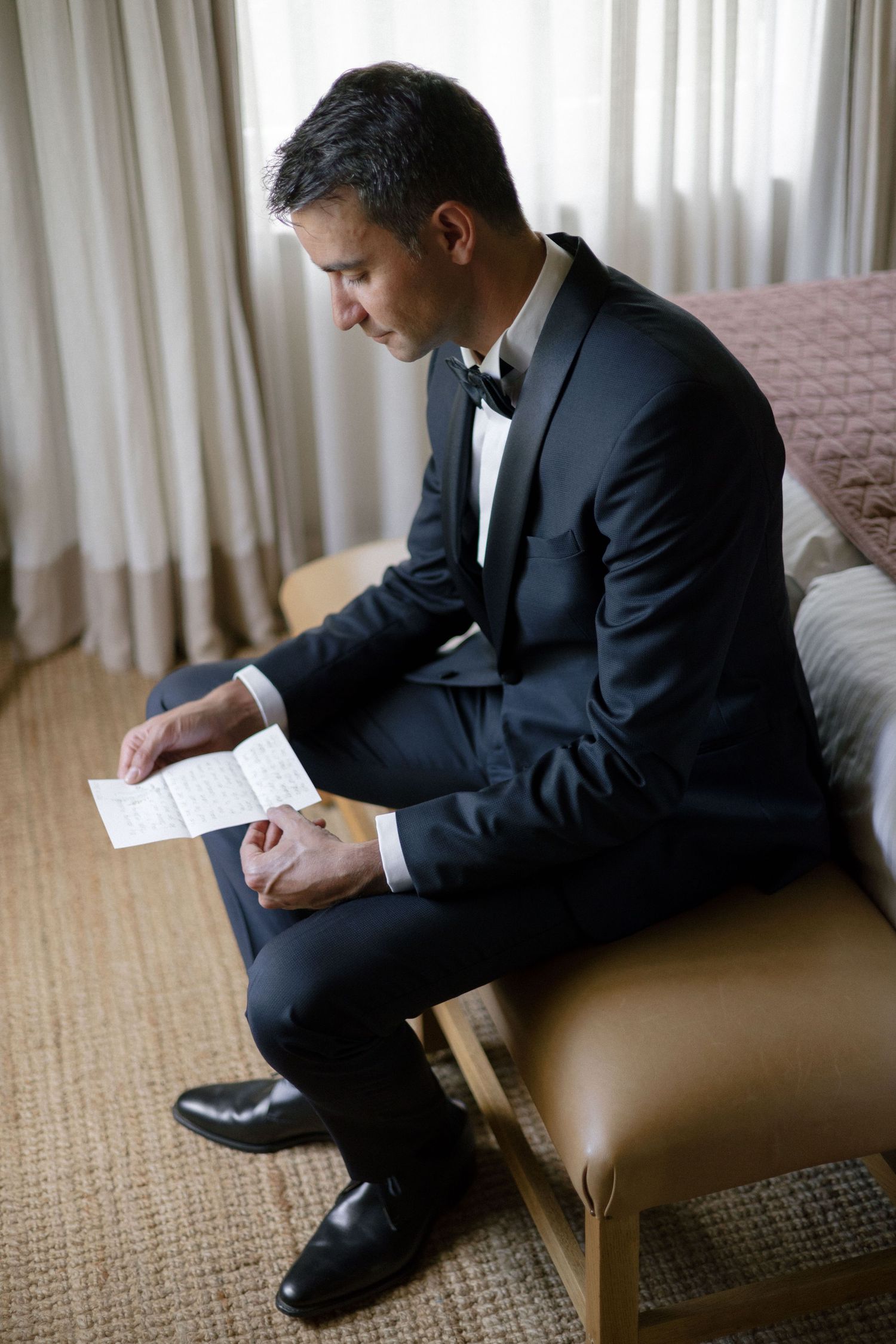 Groom in black tuxedo reading a heartfelt letter while seated near a window before his wedding.