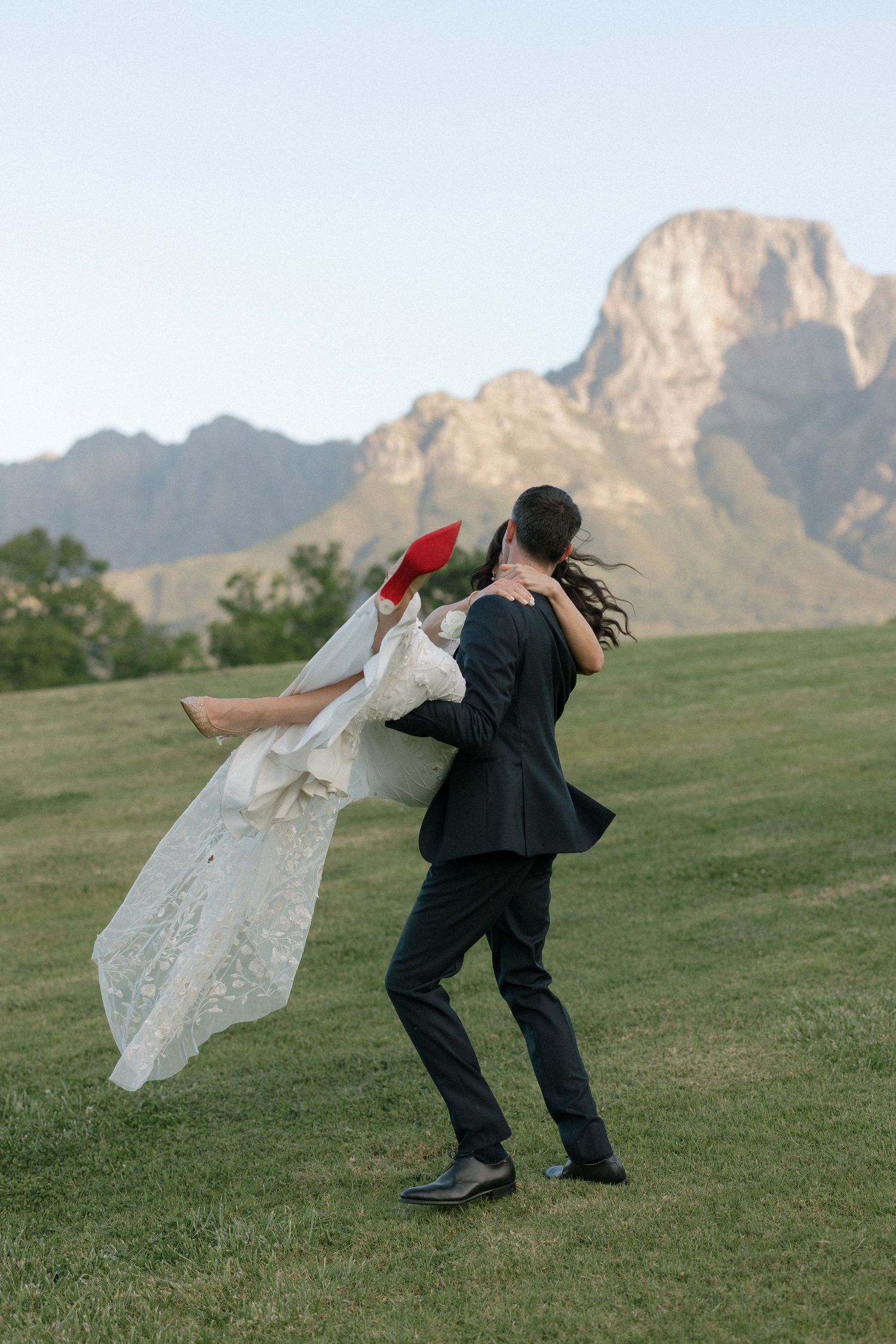Groom lifting bride in white lace dress with red heels outdoors near mountains.