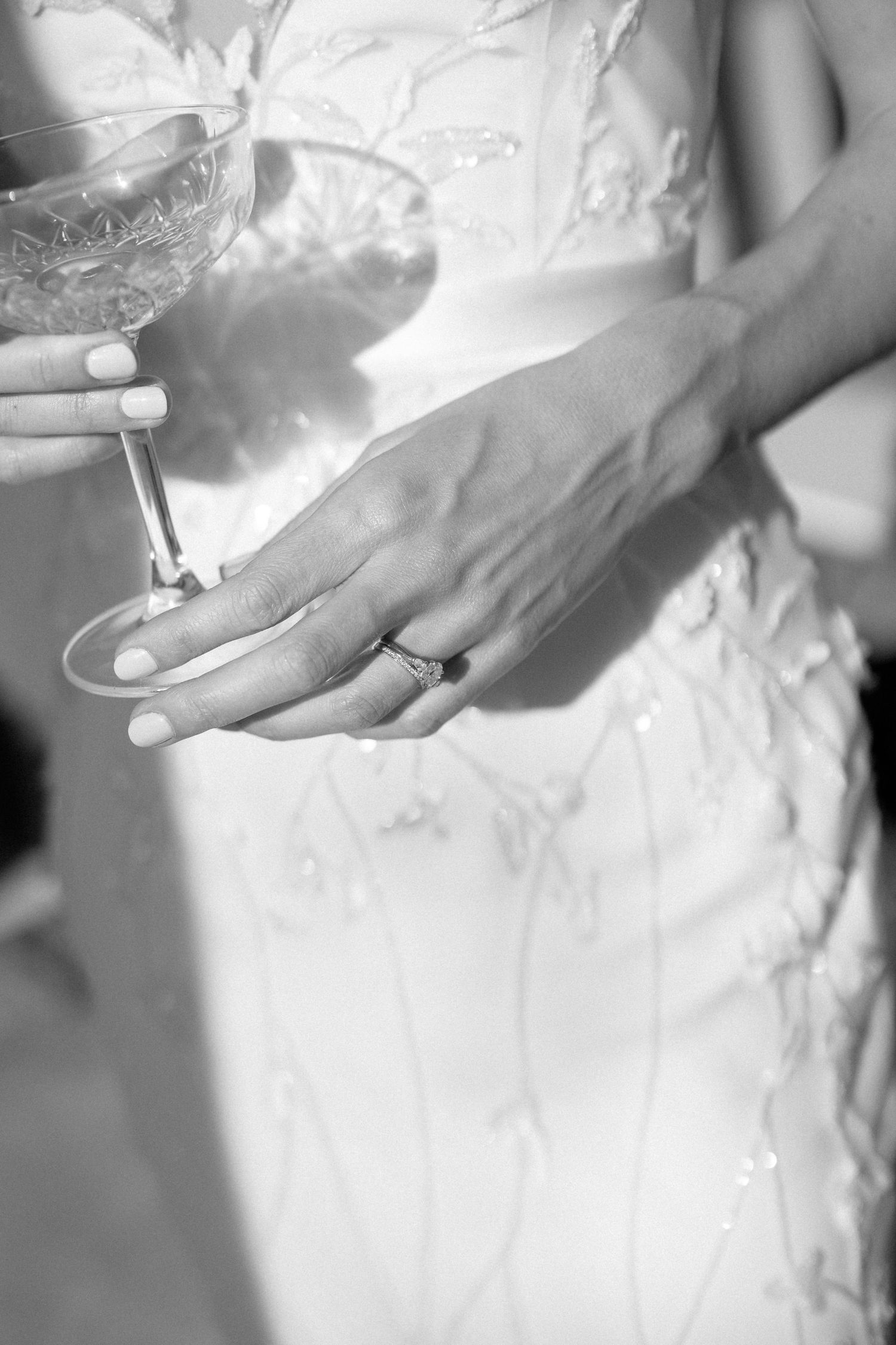 Close-up black and white photo of bride's hand with wedding ring holding crystal champagne glass.