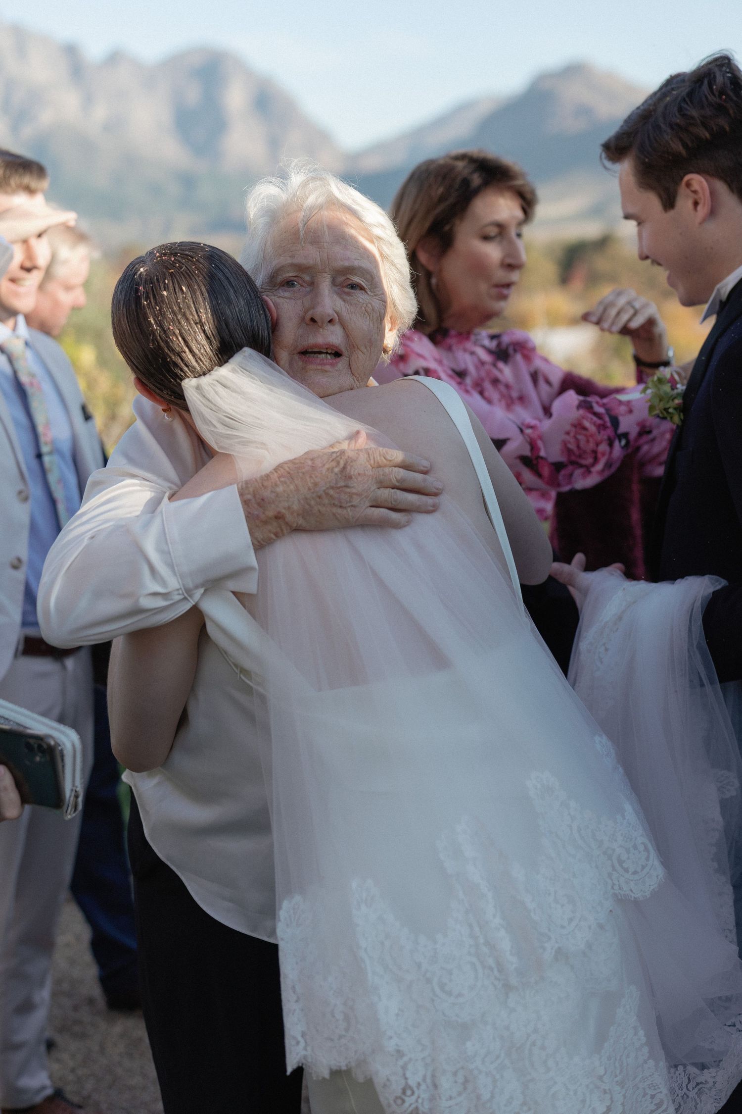 Elderly woman embraces bride in lace-trimmed veil with emotional expression during post-ceremony congratulations outdoors.