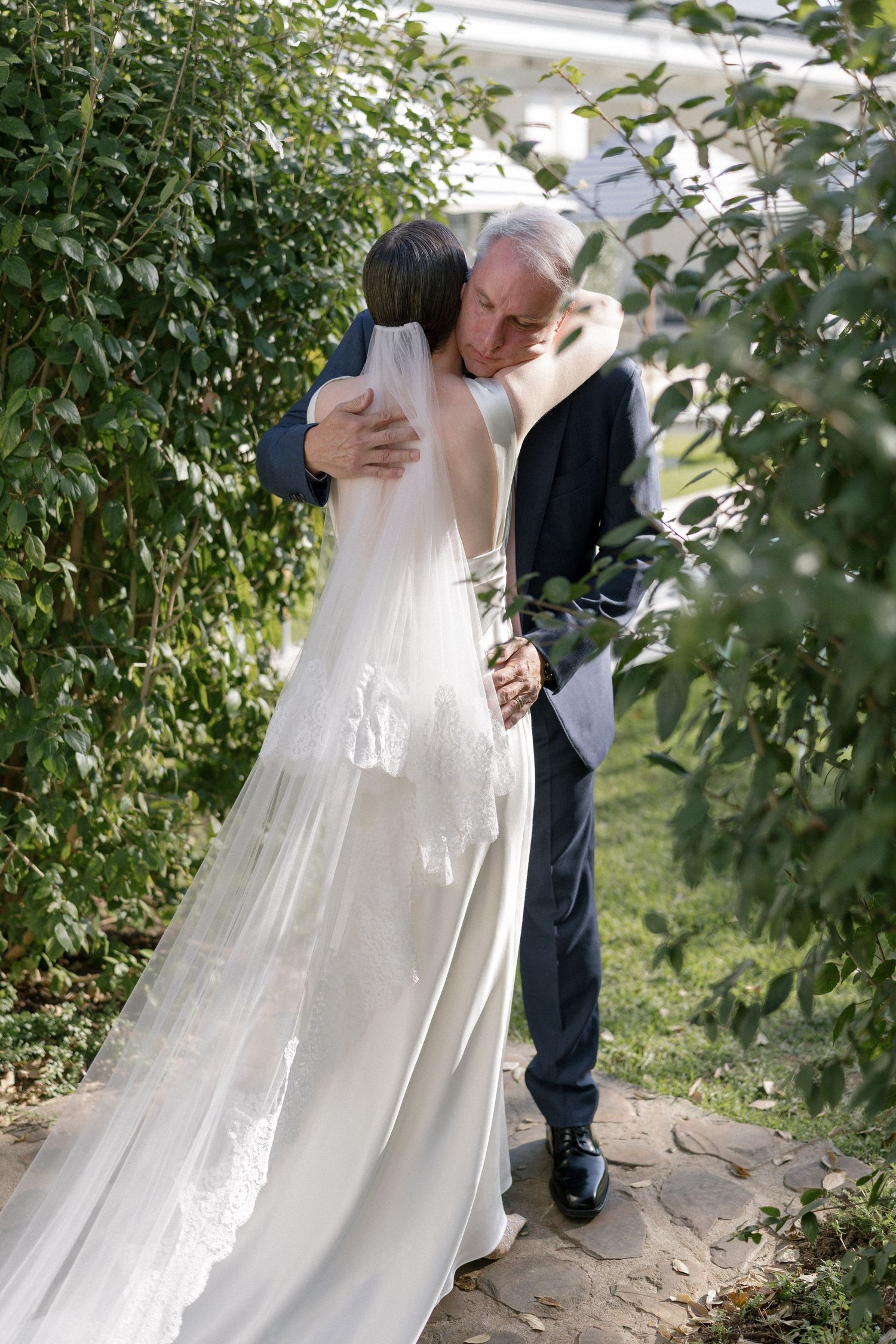 Older man embraces bride in lace-back satin gown and veil outdoors.
