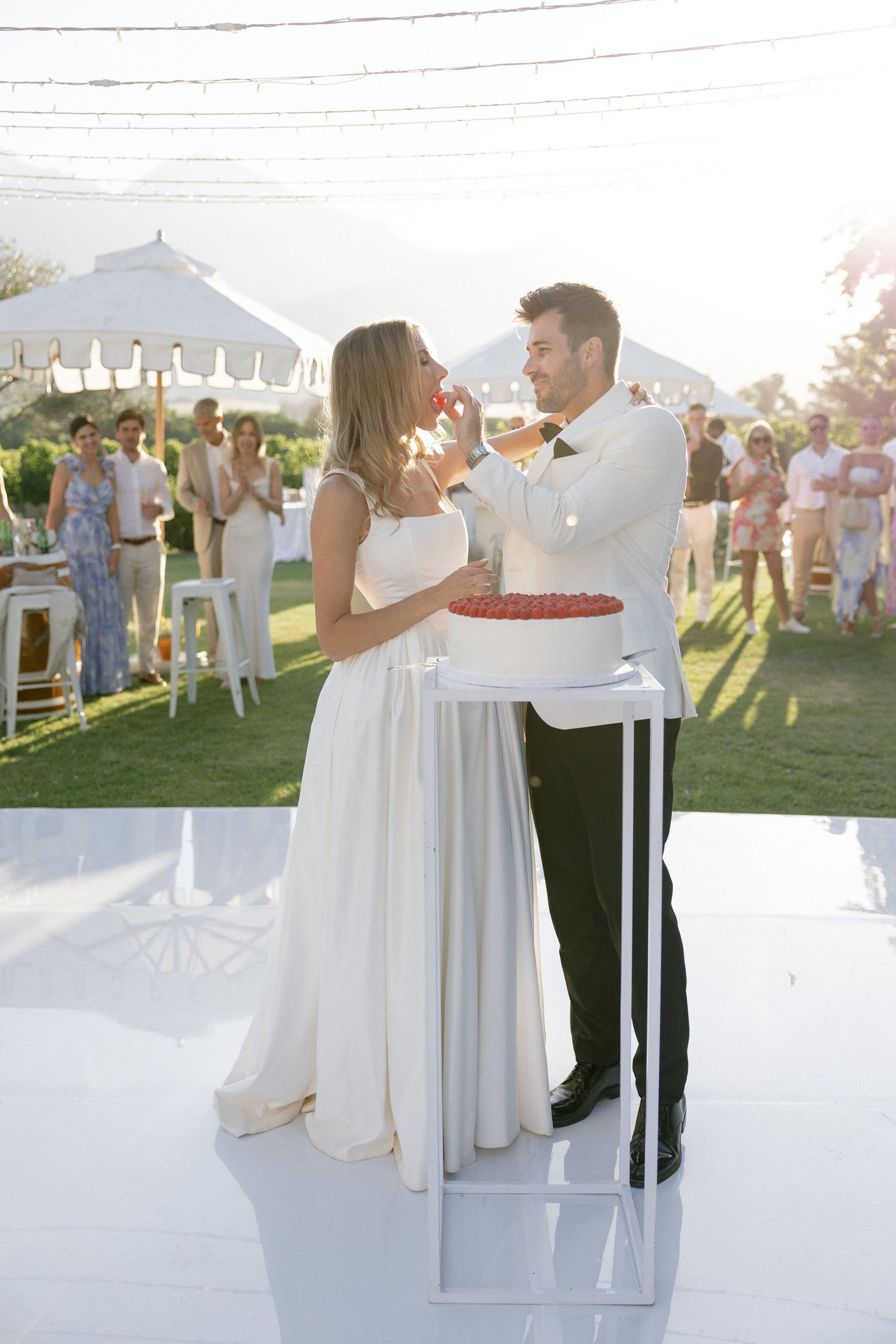 Bride and groom cutting wedding cake outdoors at sunny reception with guests celebrating in background.