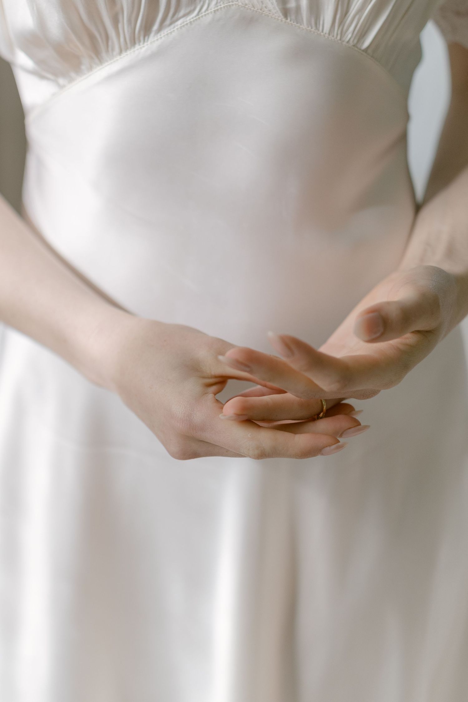 Close-up of bride's hands clasped together wearing gold ring against white satin gown.