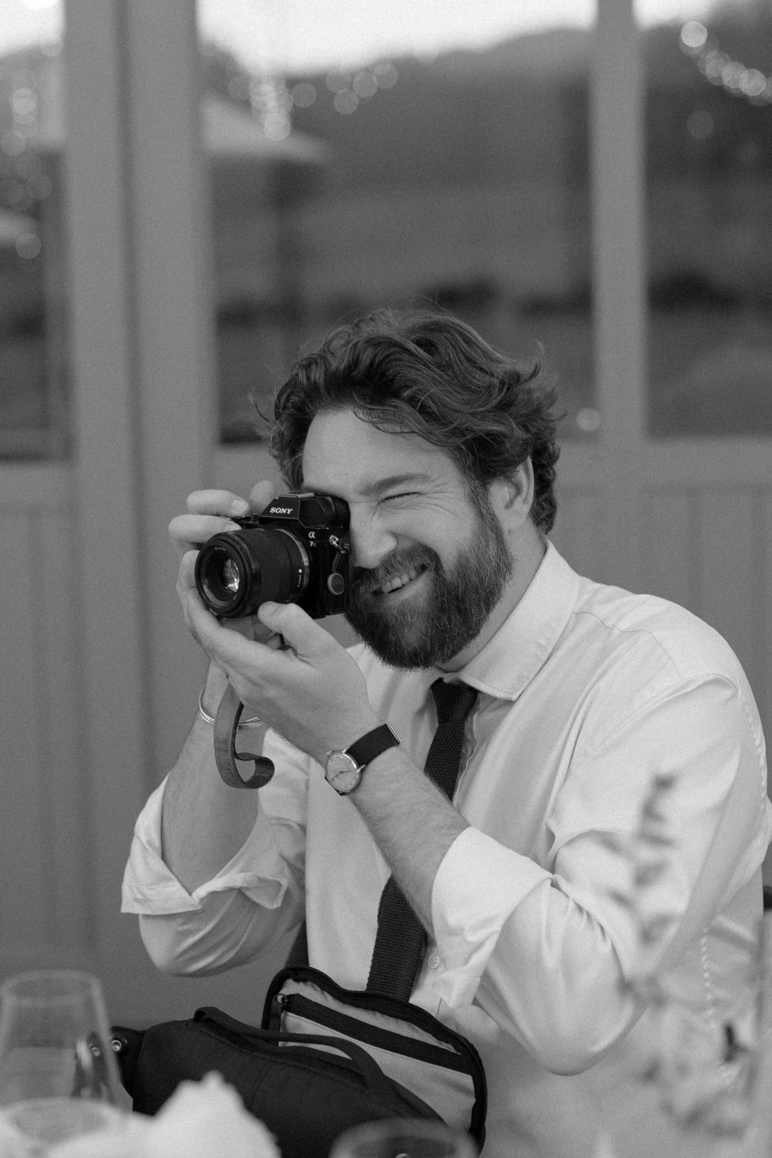 Smiling bearded male wedding guest photographing with DSLR camera at reception.