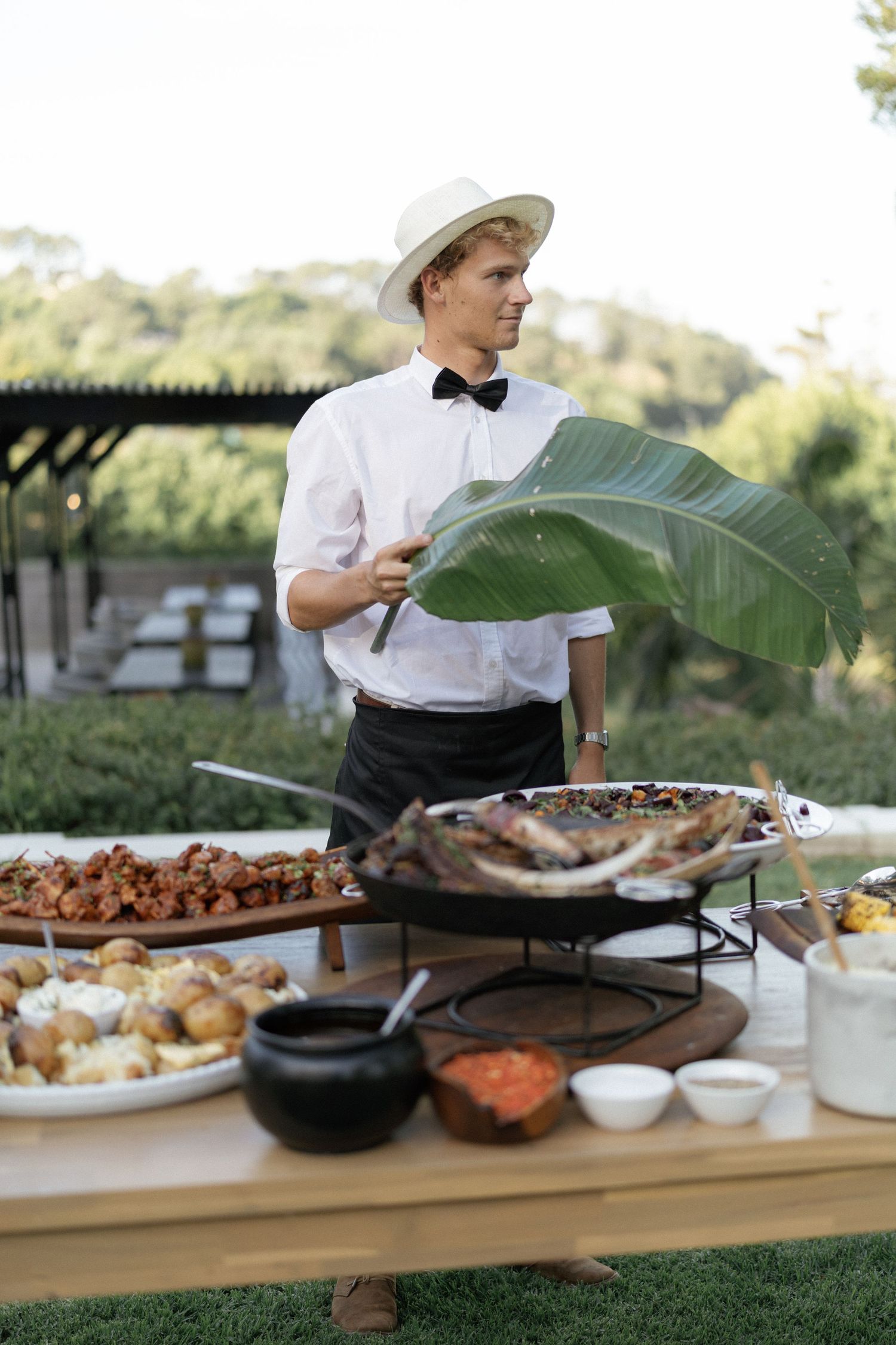 A waiter in a white shirt and hat stands behind an outdoor buffet table with various dishes at a catered event.