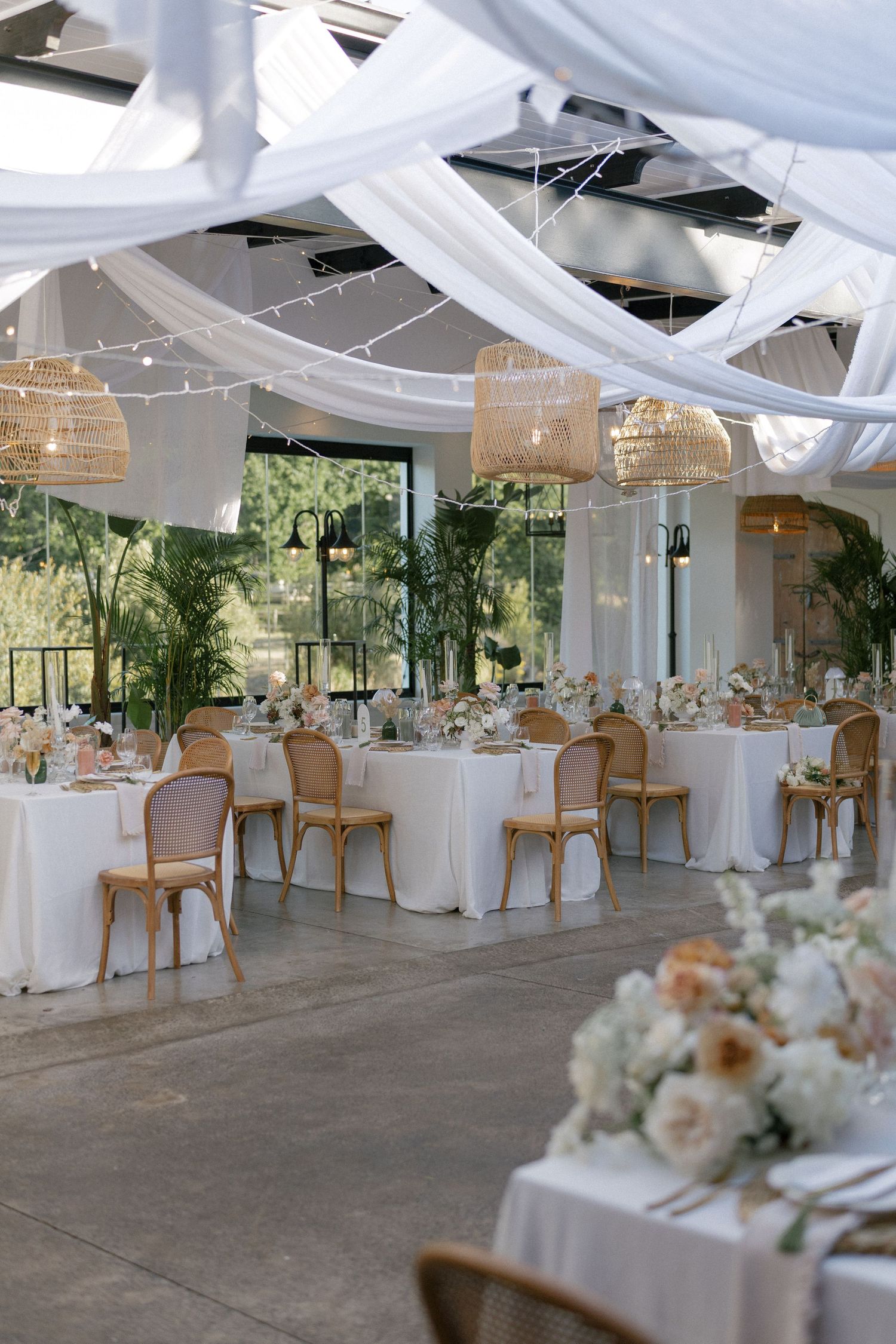 Elegant wedding reception hall with white draping, rattan pendant lights, wooden chairs, and floral centerpieces on white-clothed tables.