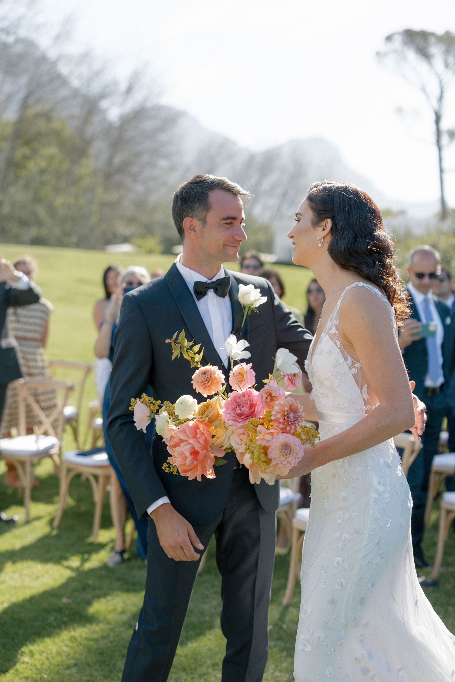 Bride and groom smiling at each other after outdoor wedding ceremony, bride holding vibrant pink and coral bouquet.
