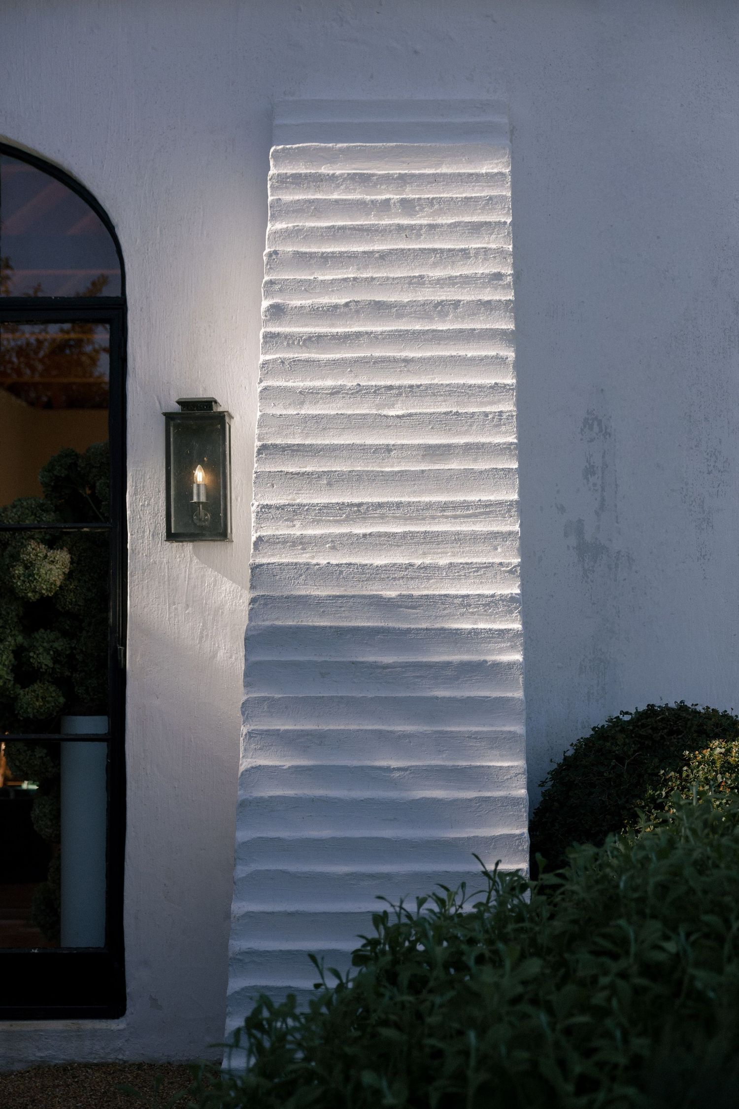 White textured wedding venue exterior wall with horizontal ridges illuminated by a vintage lantern sconce at dusk.